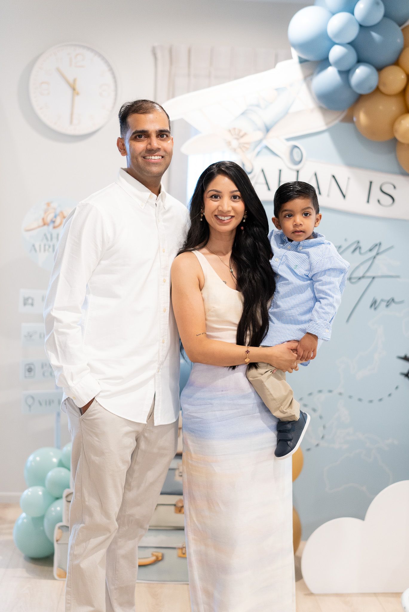 Family portrait with Ahan at his 2nd birthday celebration, standing in front of the airplane-themed party backdrop and balloons