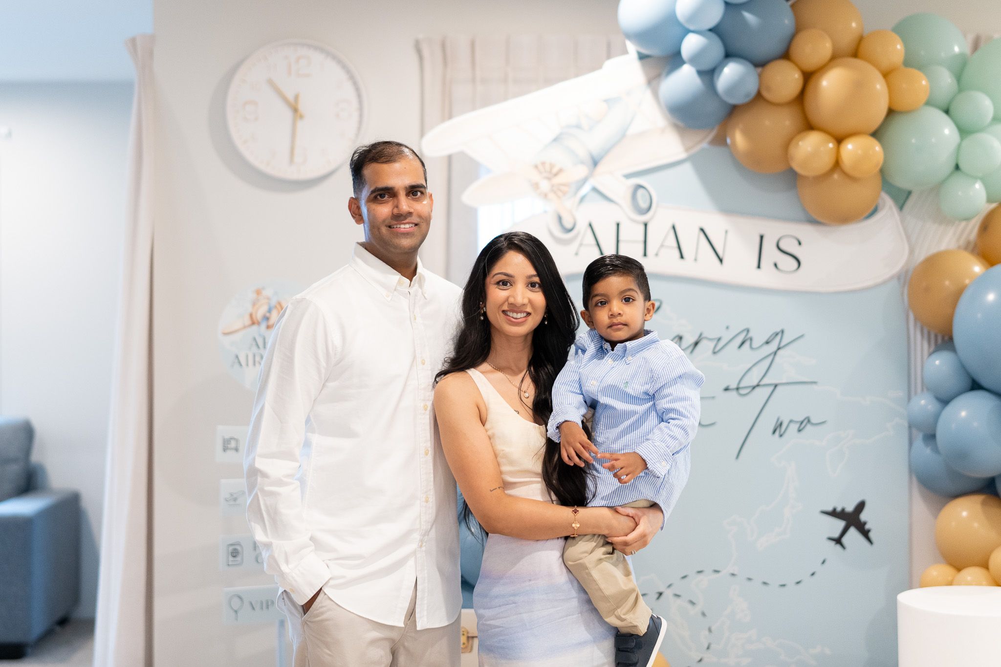 Ahan with his parents in front of the blue and gold travel-themed birthday backdrop during his 2nd birthday celebration