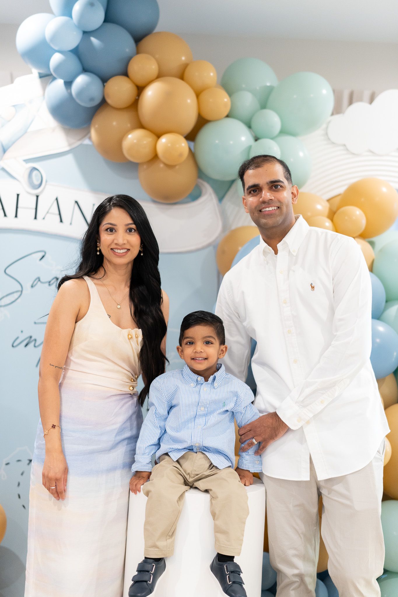 Ahan seated between his parents for a family birthday portrait, surrounded by blue, mint and gold balloon decorations
