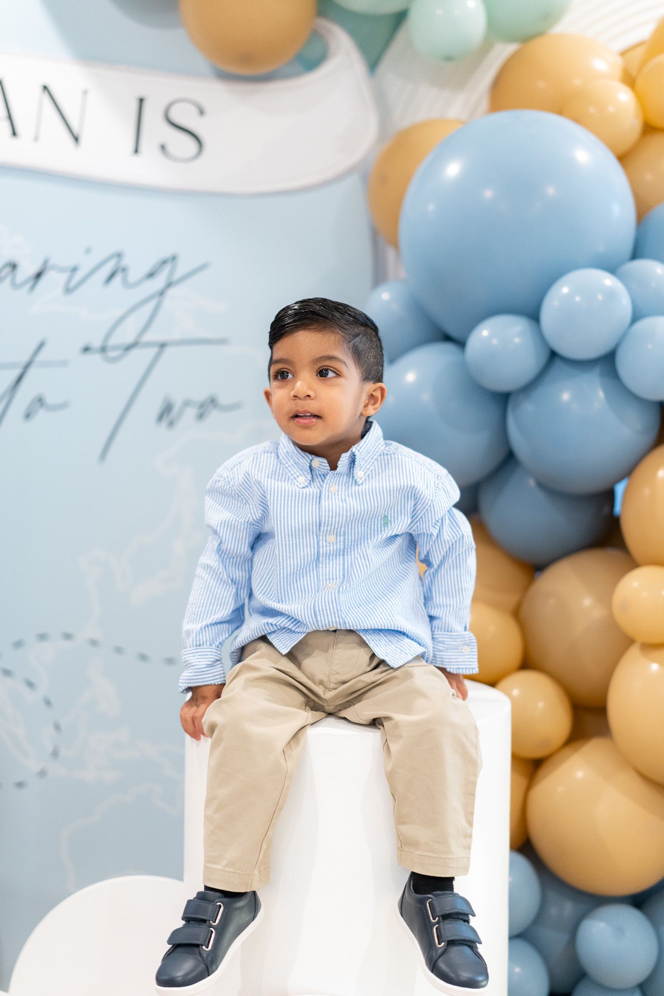 Ahan sitting on a white plinth at his 2nd birthday party, framed by blue and gold balloons and travel-themed decor