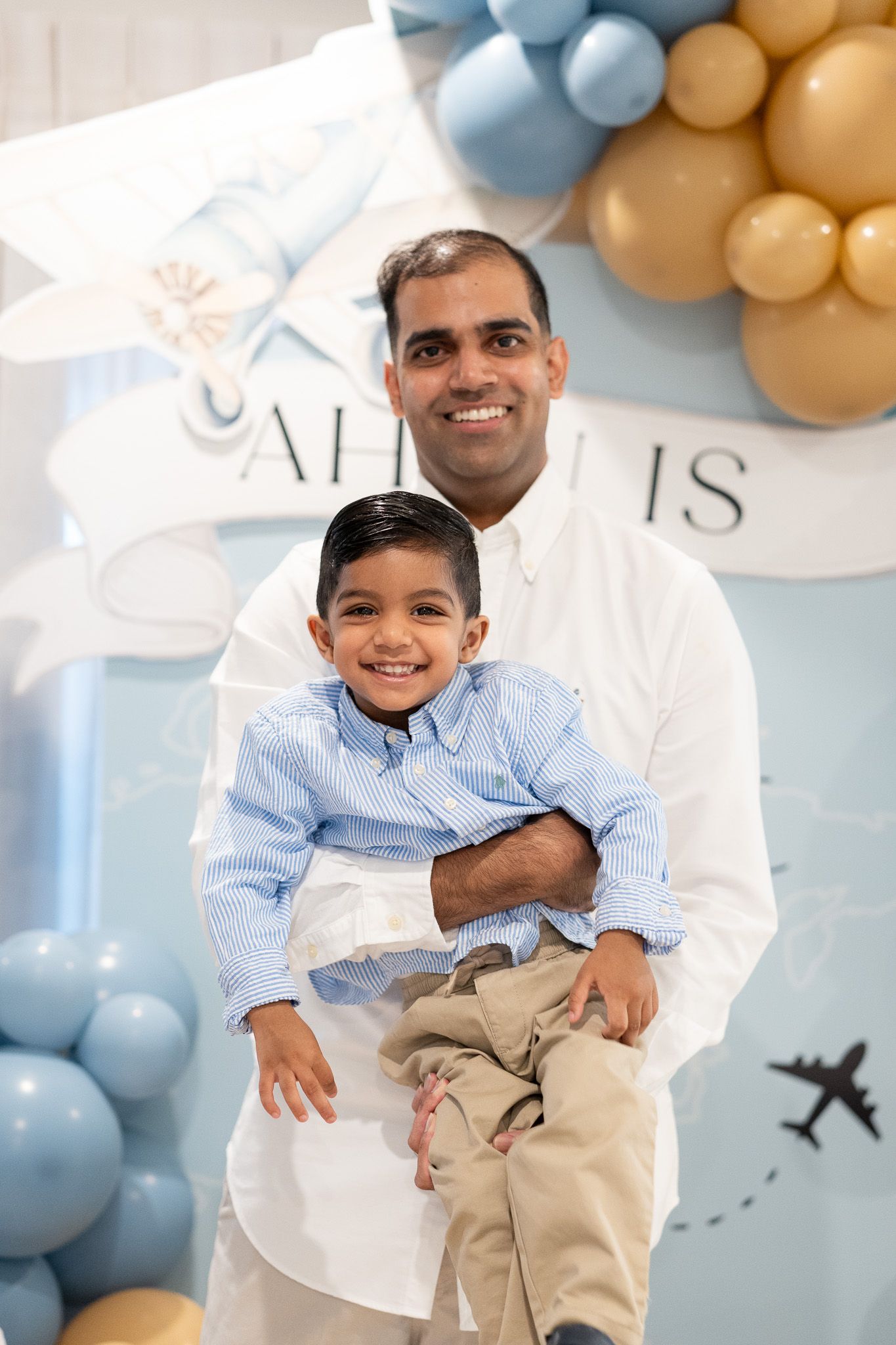 Ahan laughing while being held by his dad, with an airplane backdrop and blue and gold balloons behind them