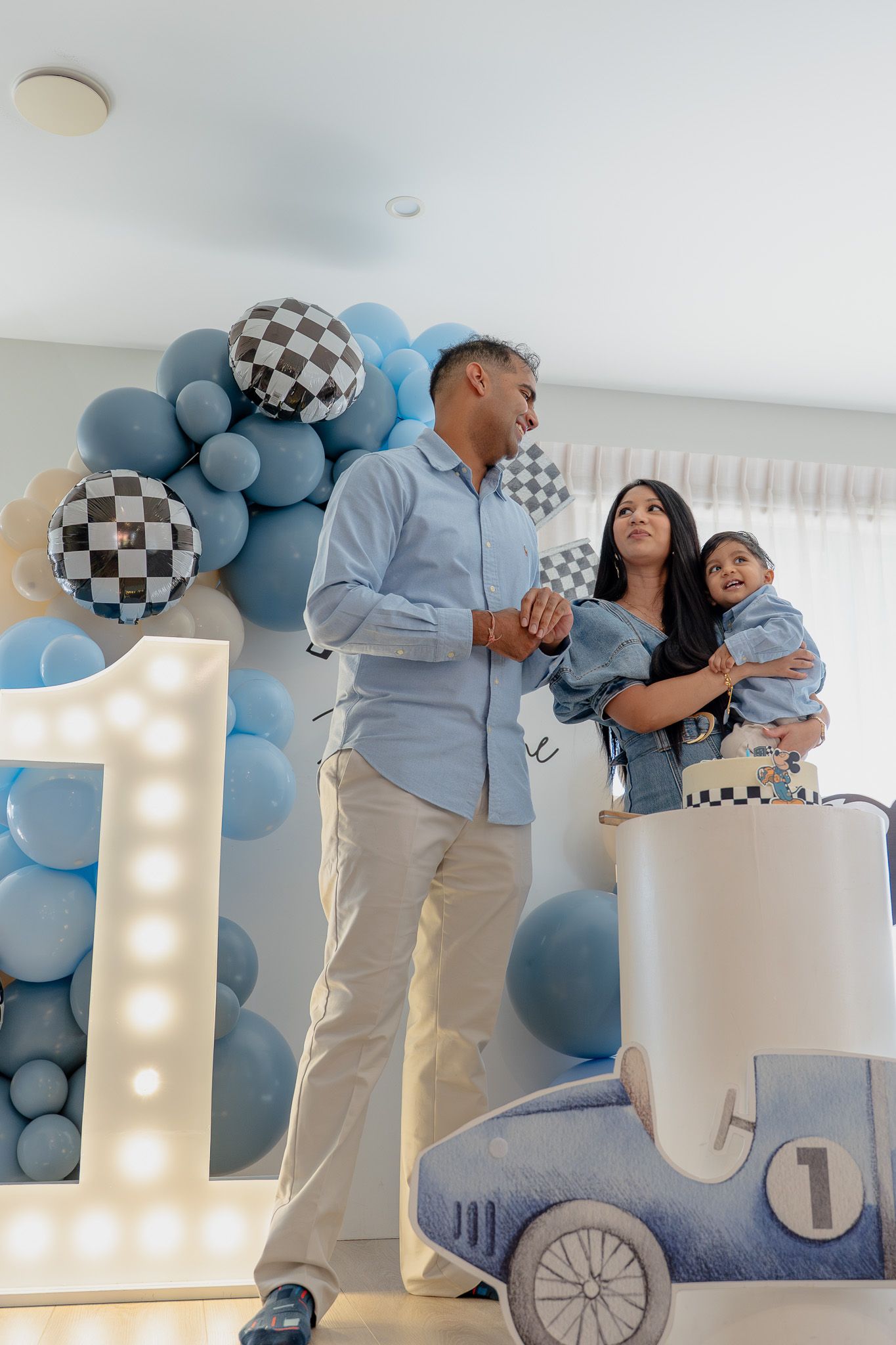 Parents and toddler celebrating together beside a racing-themed birthday cake and number 1 light prop at a kids birthday party in Auckland
