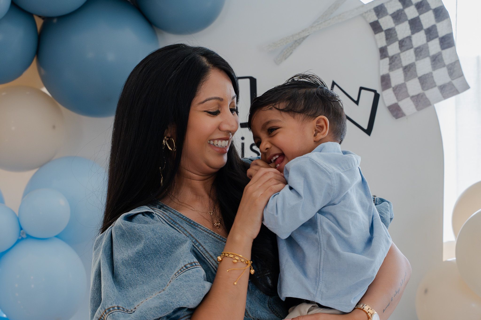 Mother laughing with her young son in front of a balloon backdrop during a joyful first birthday celebration in Auckland