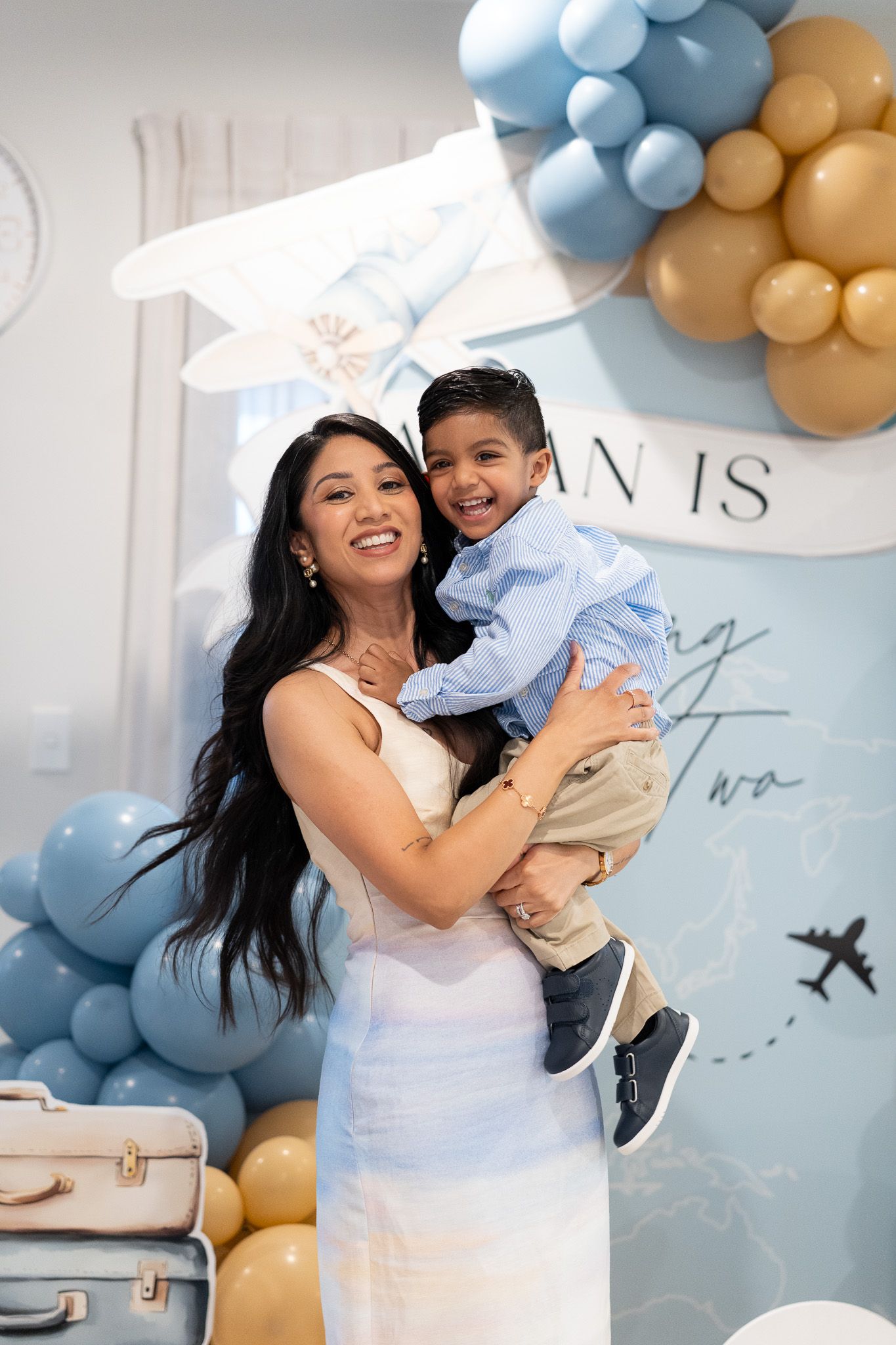 Mother holding birthday boy Ahan smiling in front of pastel travel themed balloon backdrop second birthday photography in Auckland