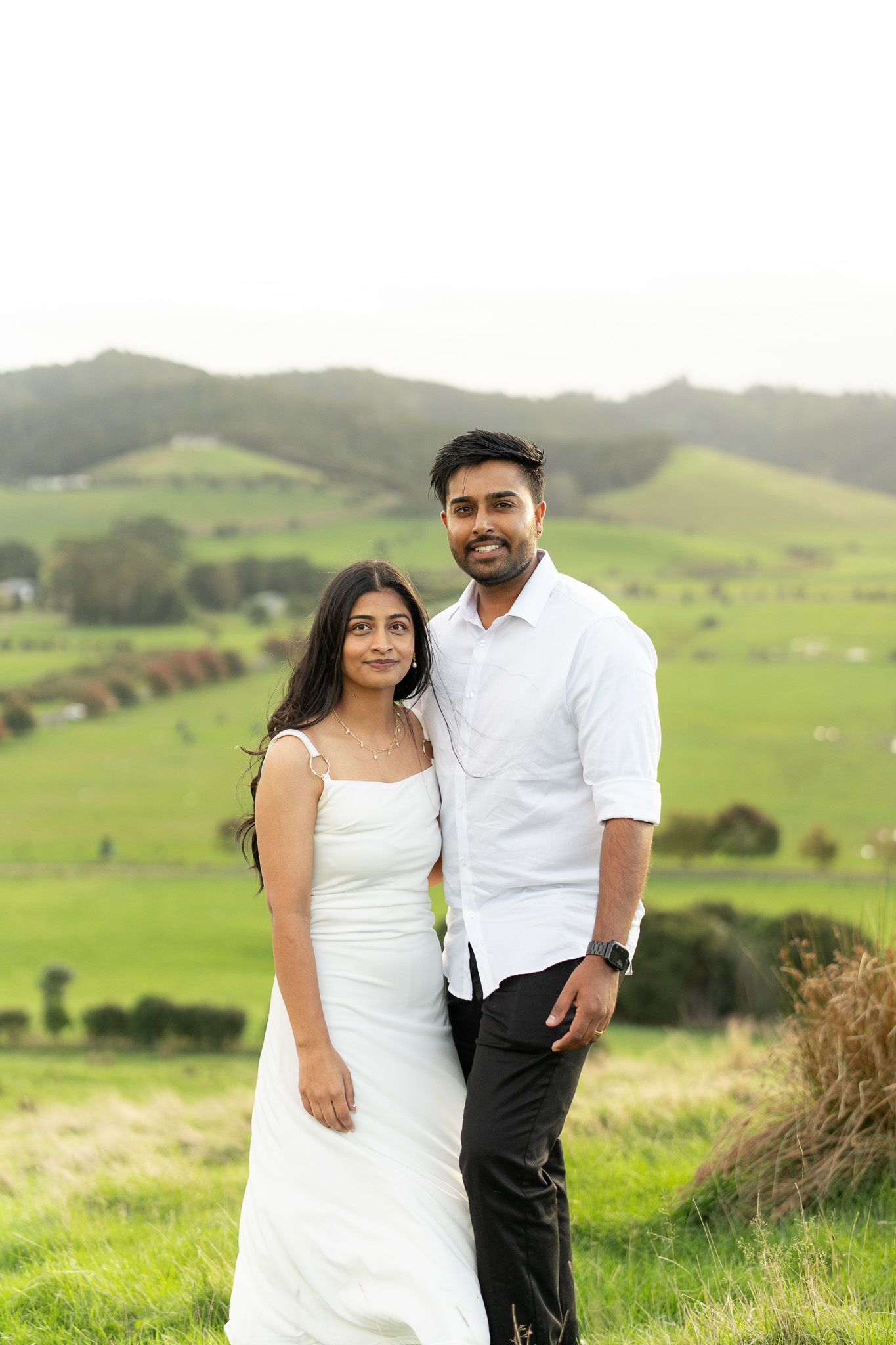 Amish and Komal standing together on a green hillside during a relaxed couples photography session with soft countryside views