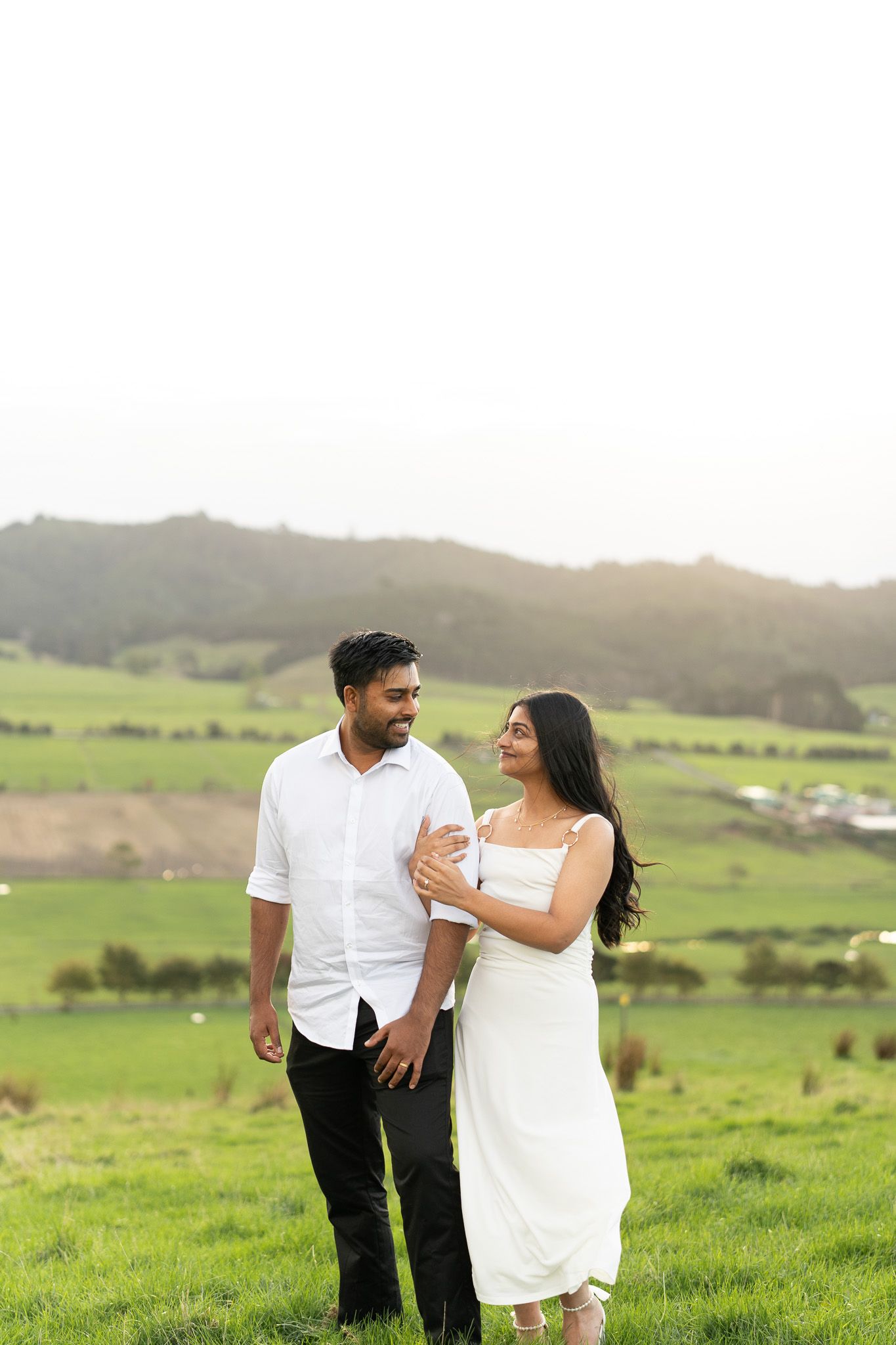 Komal holding Amish’s arm as they walk through long grass during a relaxed couples photography session in Auckland