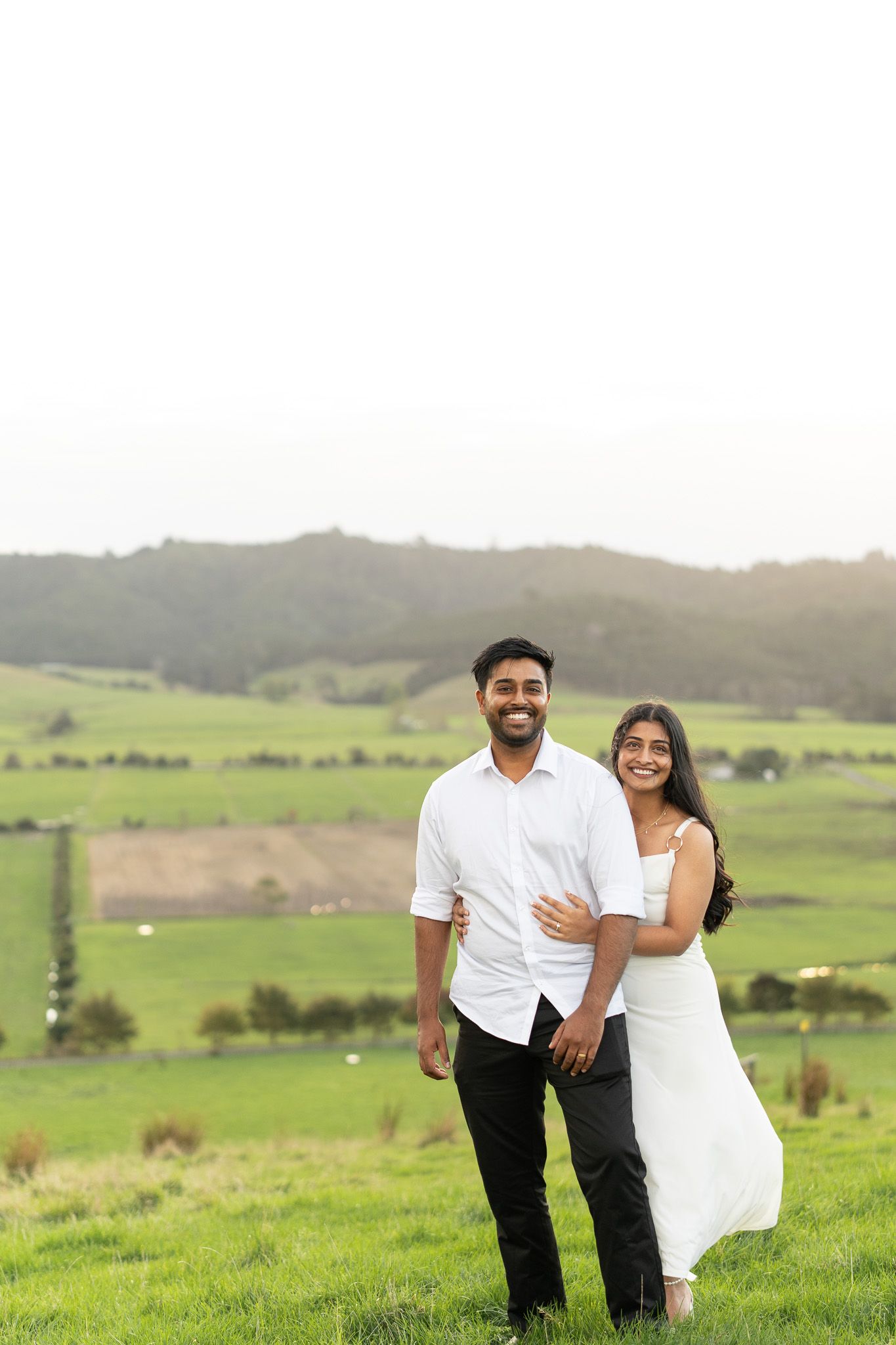 Amish and Komal smiling together on a green hillside with soft cloudy sunset light during their couples photoshoot