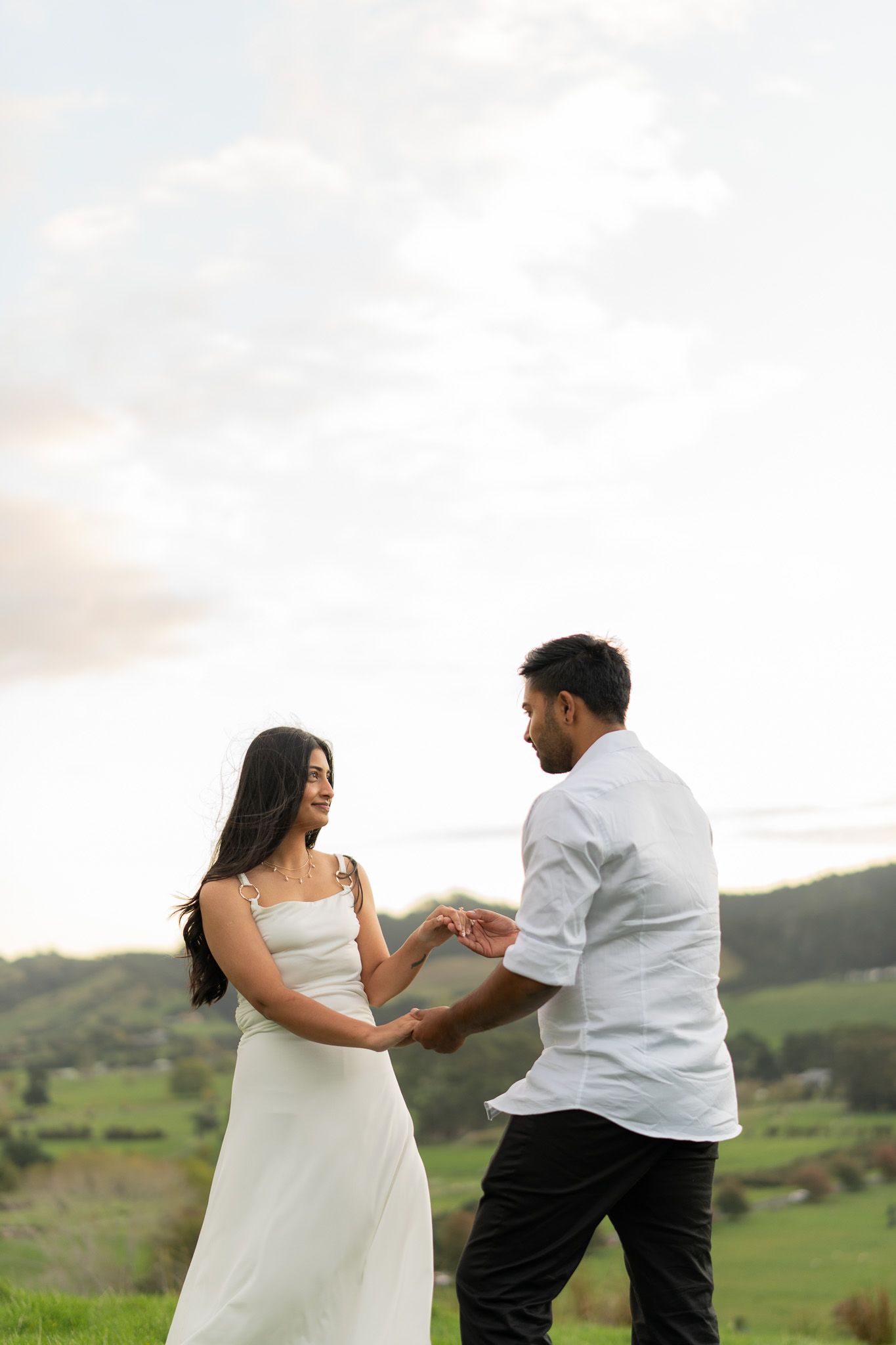 Romantic couples photography in Auckland with couple dancing together on hilltop during golden hour sunset with soft sky backdrop