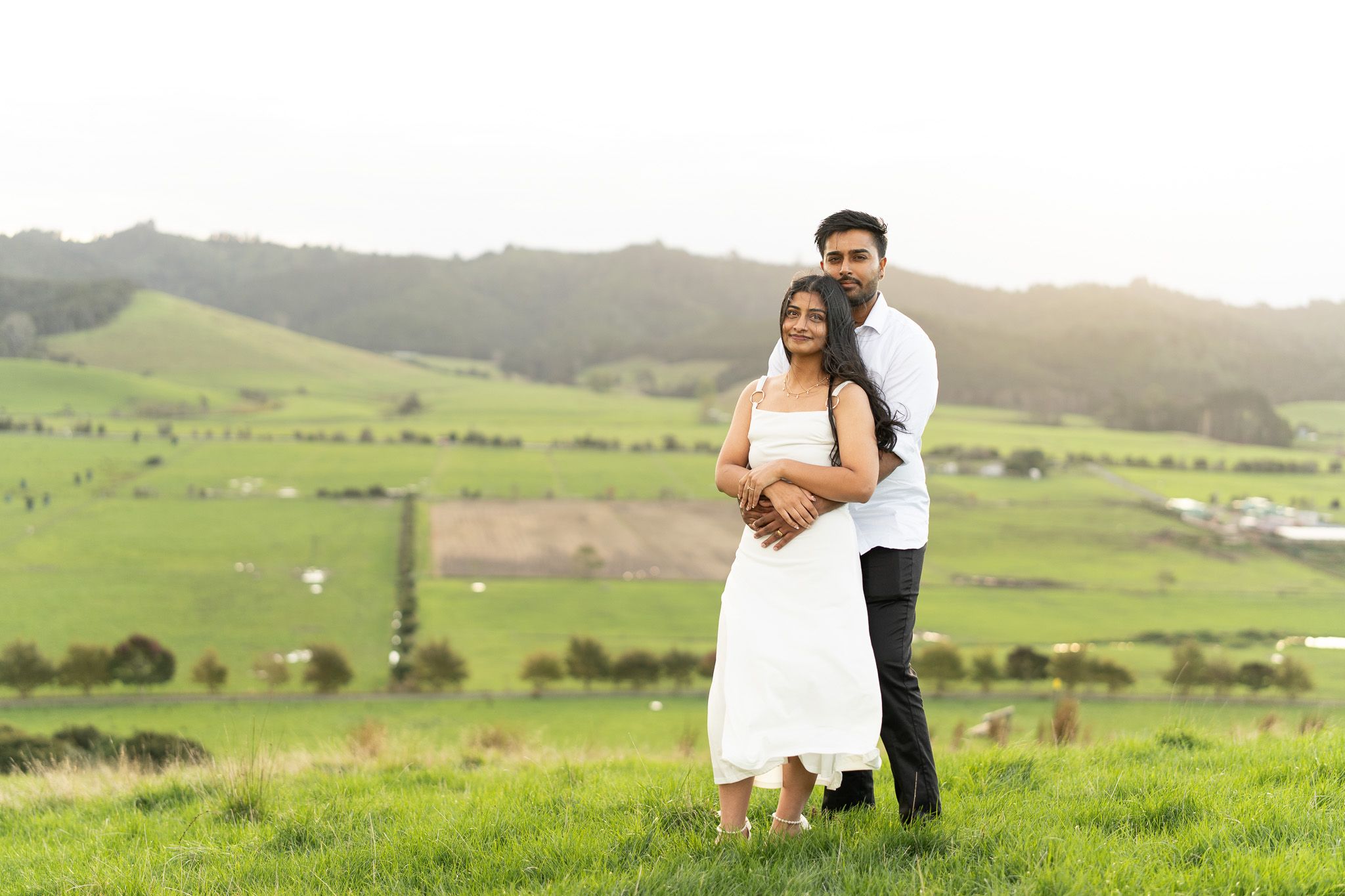 Romantic couples photography at sunset in Auckland with couple embracing on grassy hill overlooking scenic countryside landscape