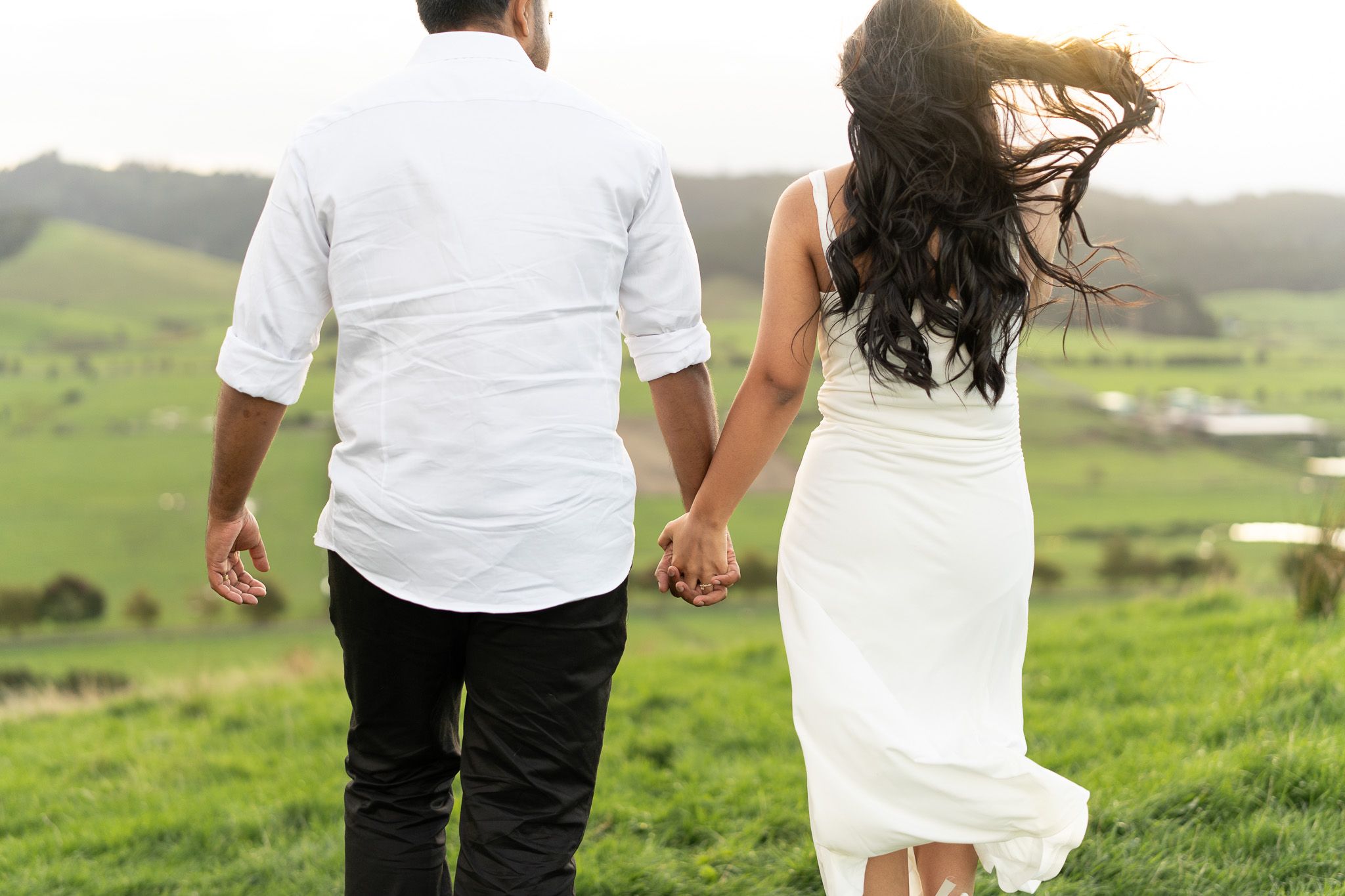 Couples walking hand in hand at sunset with wind flowing dress on hilltop captured in natural couples photography in Auckland