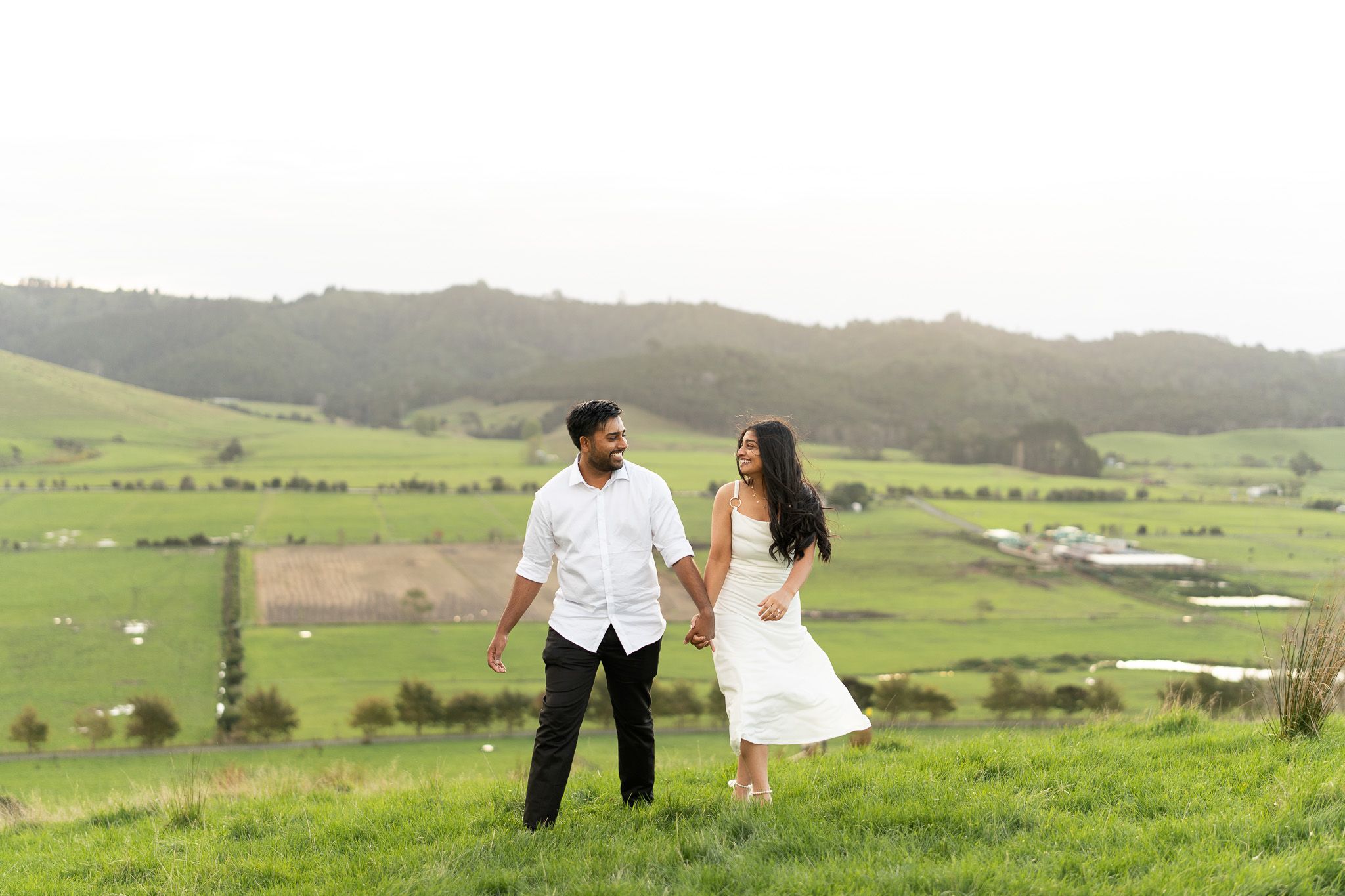 Playful couples photography in Auckland with couple walking and laughing together on green hillside during golden hour sunset