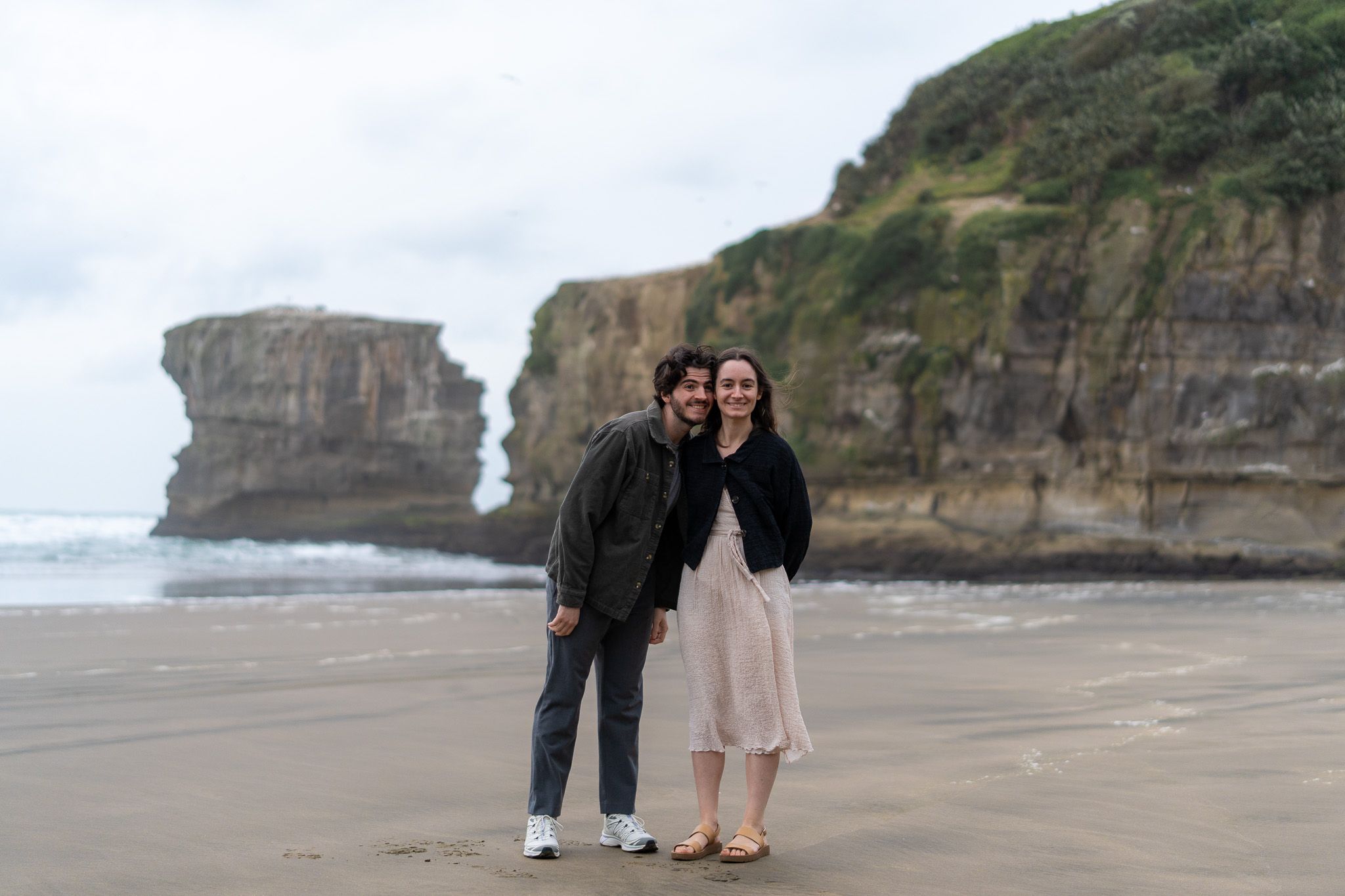 Antoine and Cyrielle smiling together on Muriwai Beach in Auckland, with rugged coastal cliffs and sea stack behind them during a moody couples photoshoot