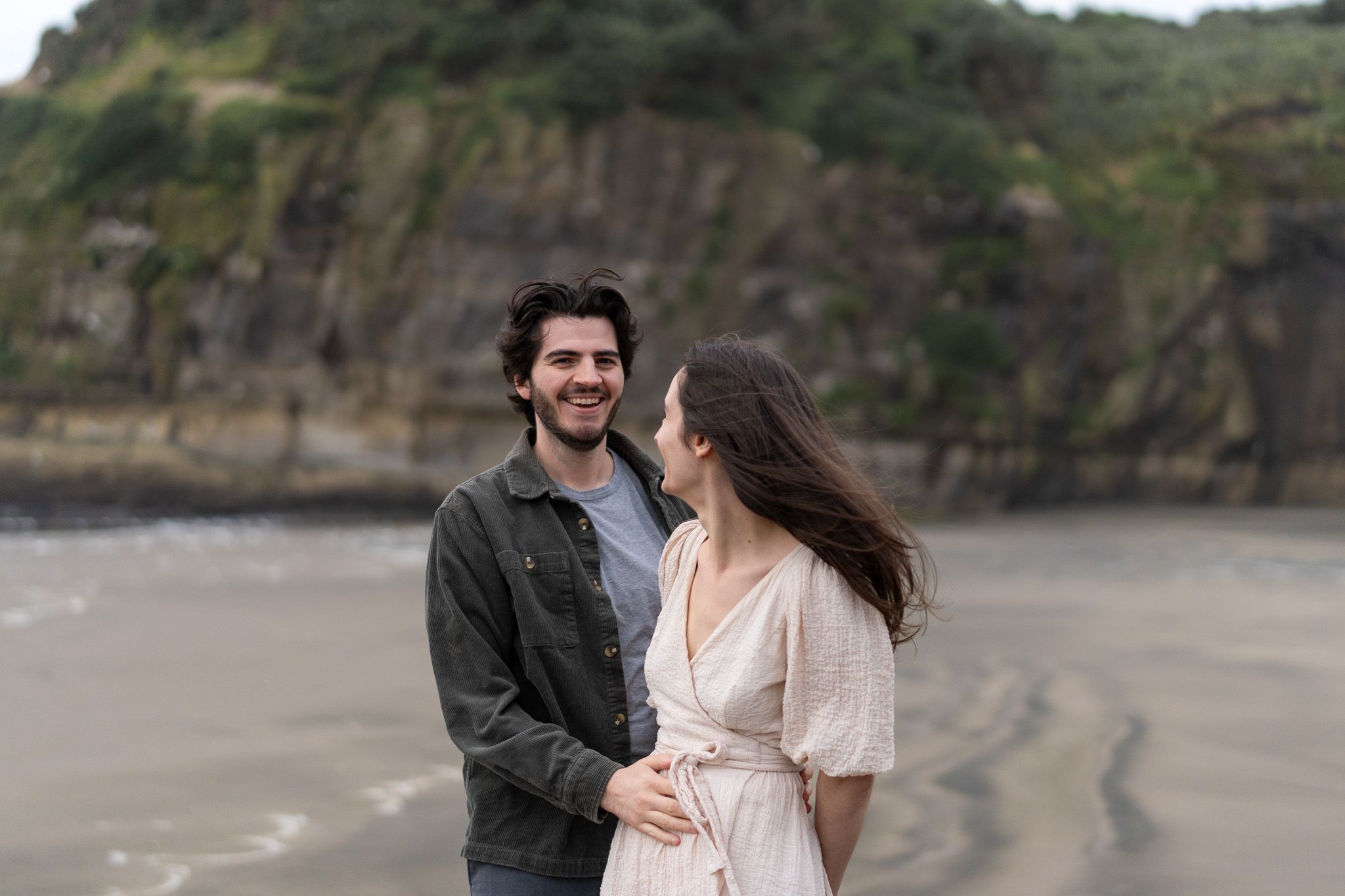 Antoine laughing while holding Cyrielle close on Muriwai Beach, with soft coastal cliffs behind them during a relaxed couples photography session