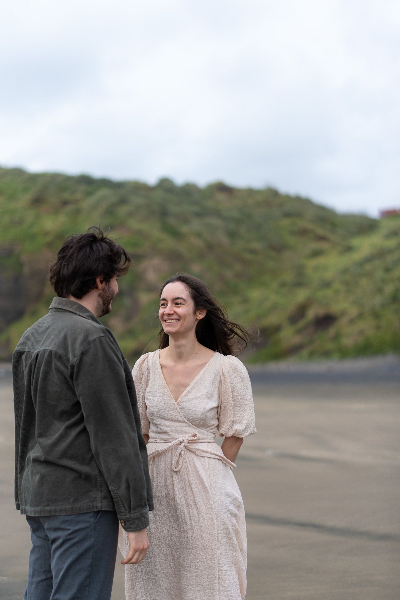 Cyrielle smiling at Antoine on Muriwai Beach in Auckland, captured against soft green cliffs and wet sand during a romantic couples photoshoot