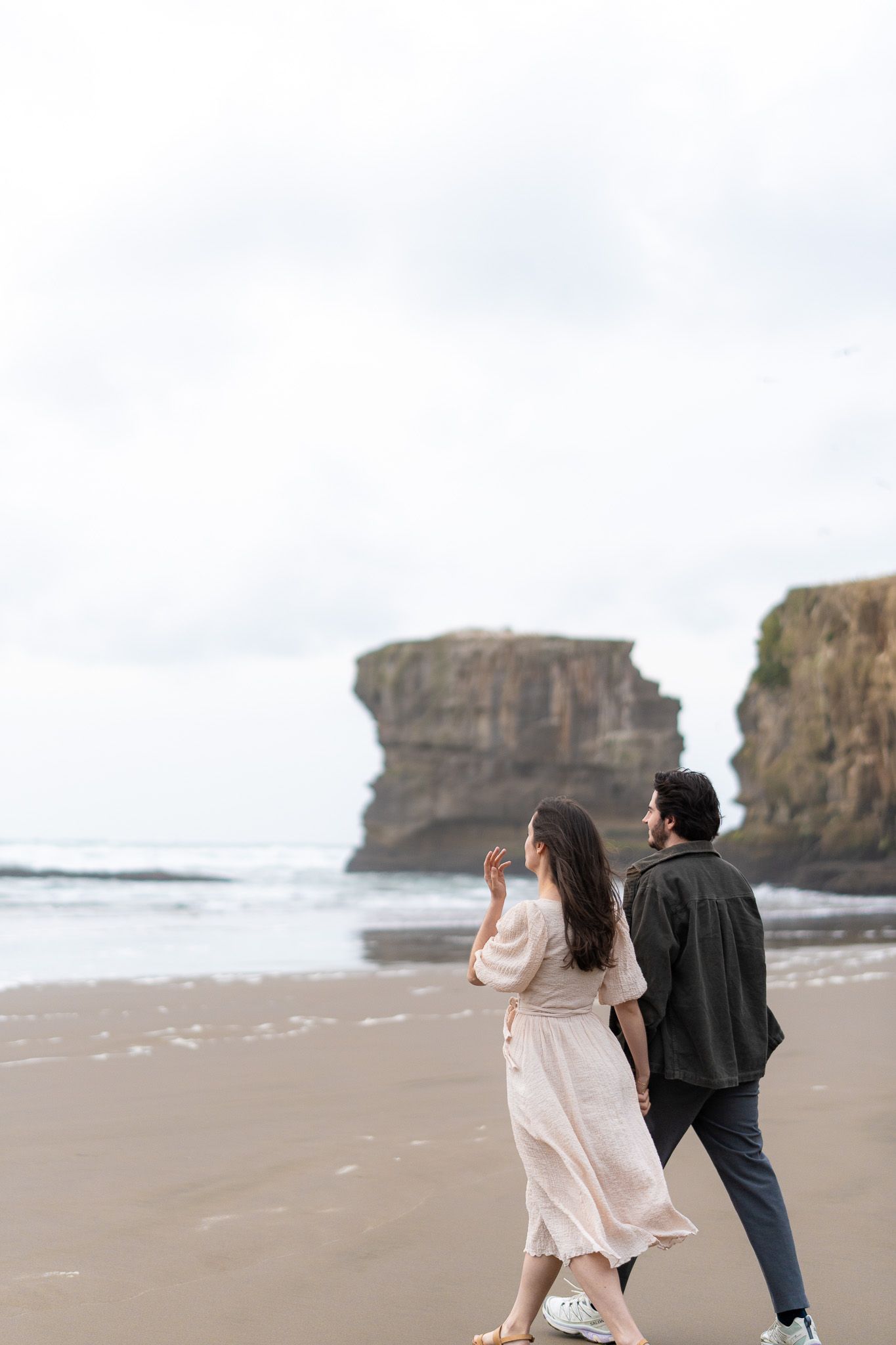 Antoine and Cyrielle walking hand in hand along Muriwai Beach, with dramatic cliffs and ocean waves creating a moody coastal couple portrait