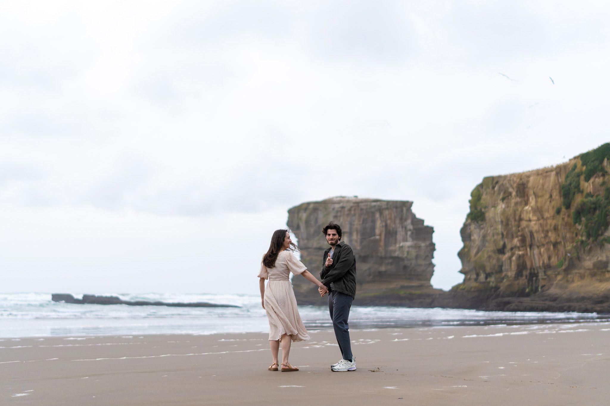 Antoine and Cyrielle holding hands on Muriwai Beach in Auckland, framed by a dramatic sea stack and cloudy sky during their couples photoshoot