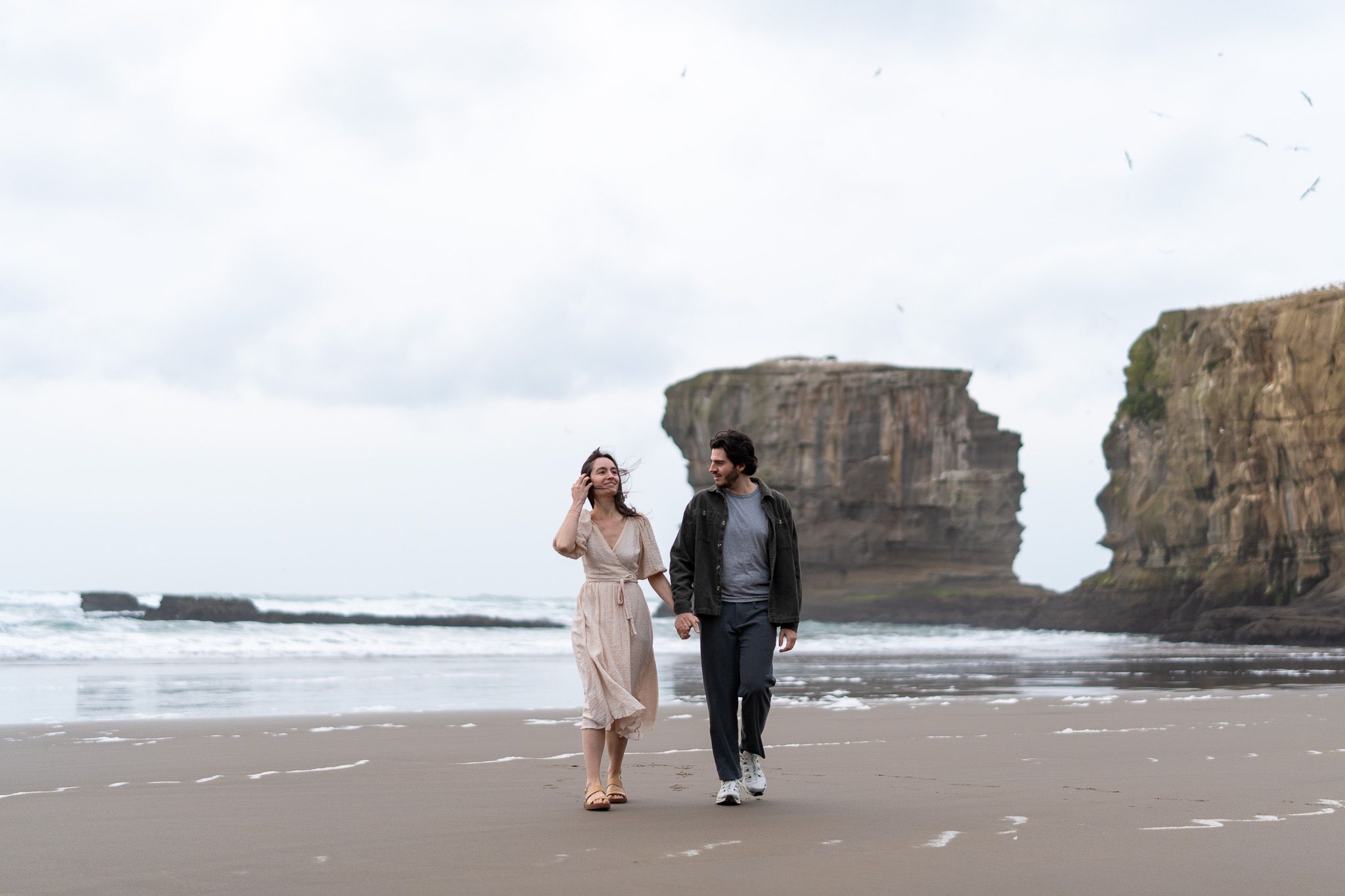 Antoine and Cyrielle walking together along the shoreline at Muriwai Beach, with waves, cliffs and seabirds behind them in Auckland