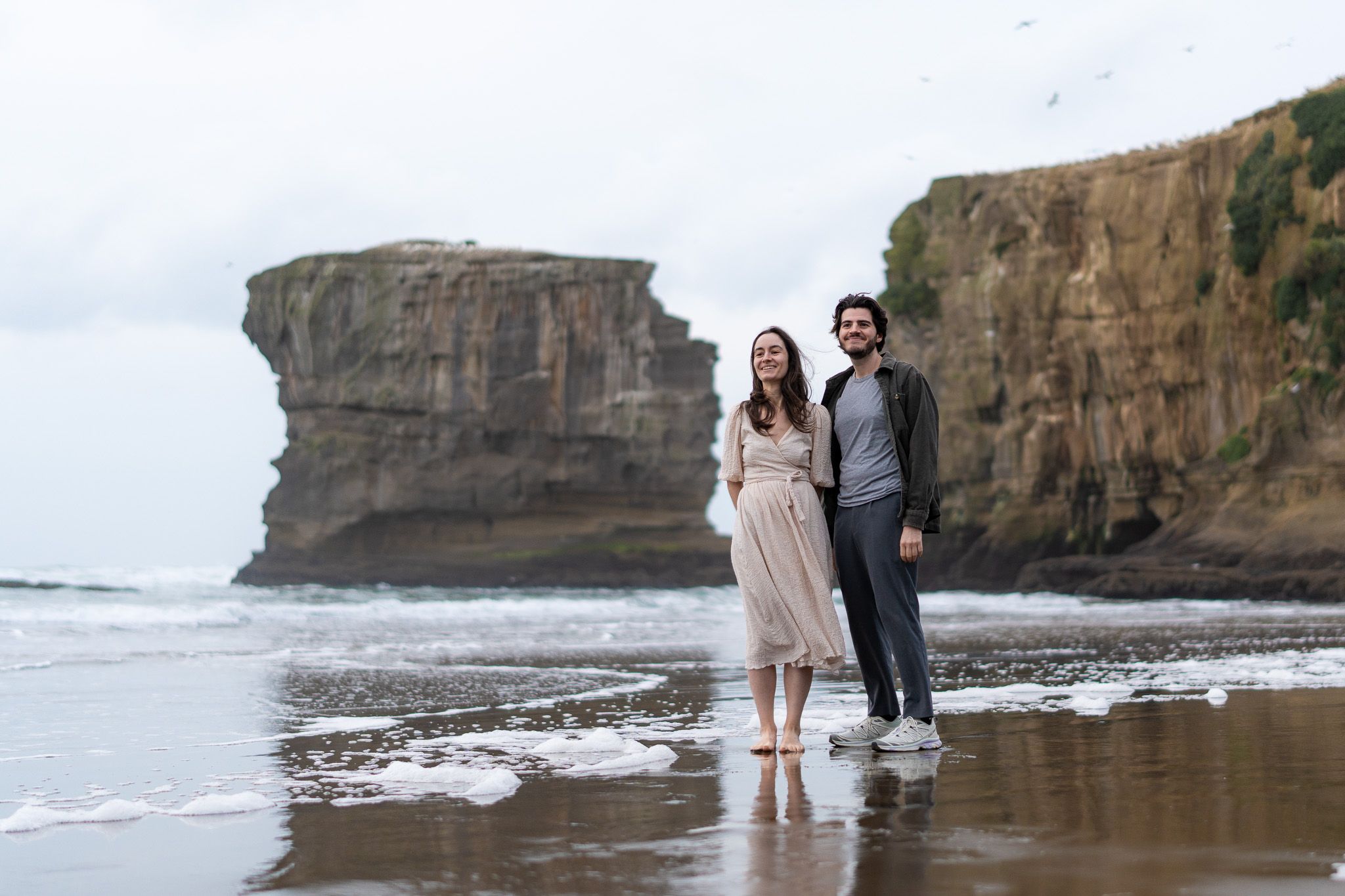 Antoine and Cyrielle standing near the water at Muriwai Beach in Auckland, with sea foam, cliff reflections and a dramatic coastal backdrop