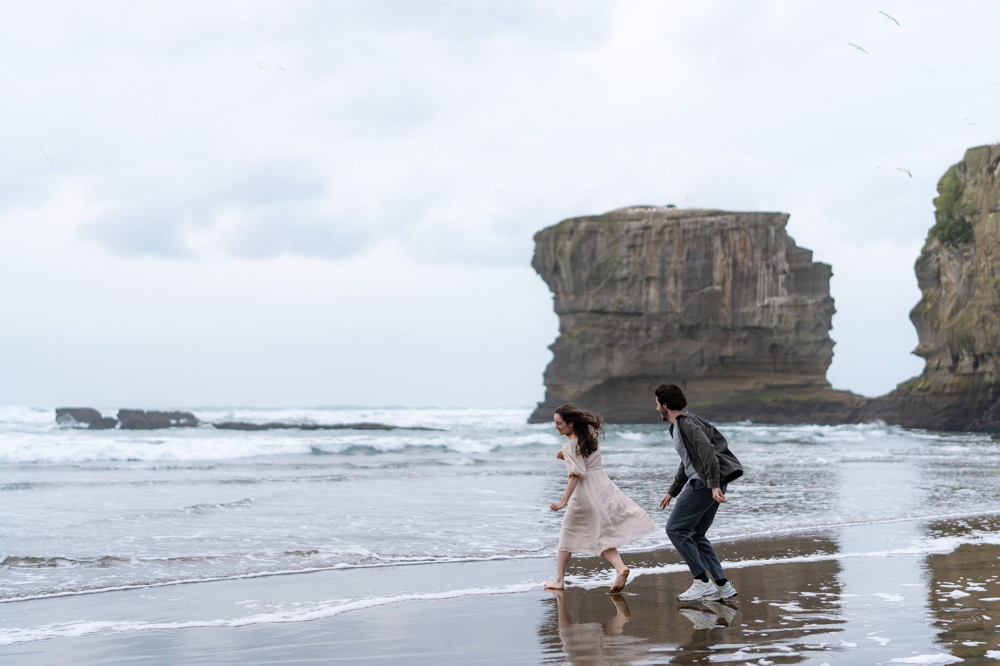 Antoine and Cyrielle running playfully along Muriwai Beach, with ocean waves and rugged cliffs behind them during a natural couples session