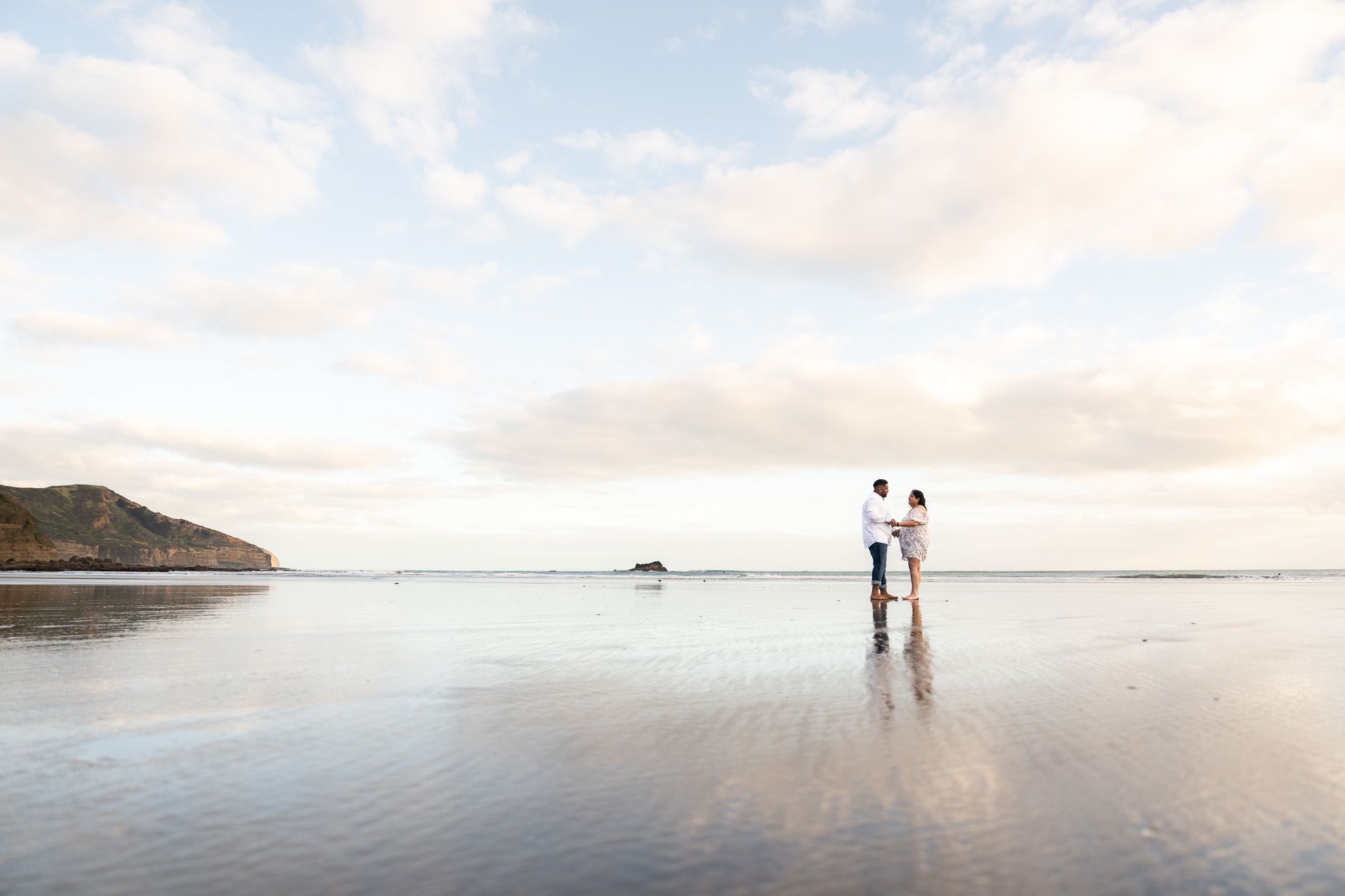 Romantic couple standing hand-in-hand on the smooth reflective shore of Muriwai Beach in Auckland, captured during golden hour photography