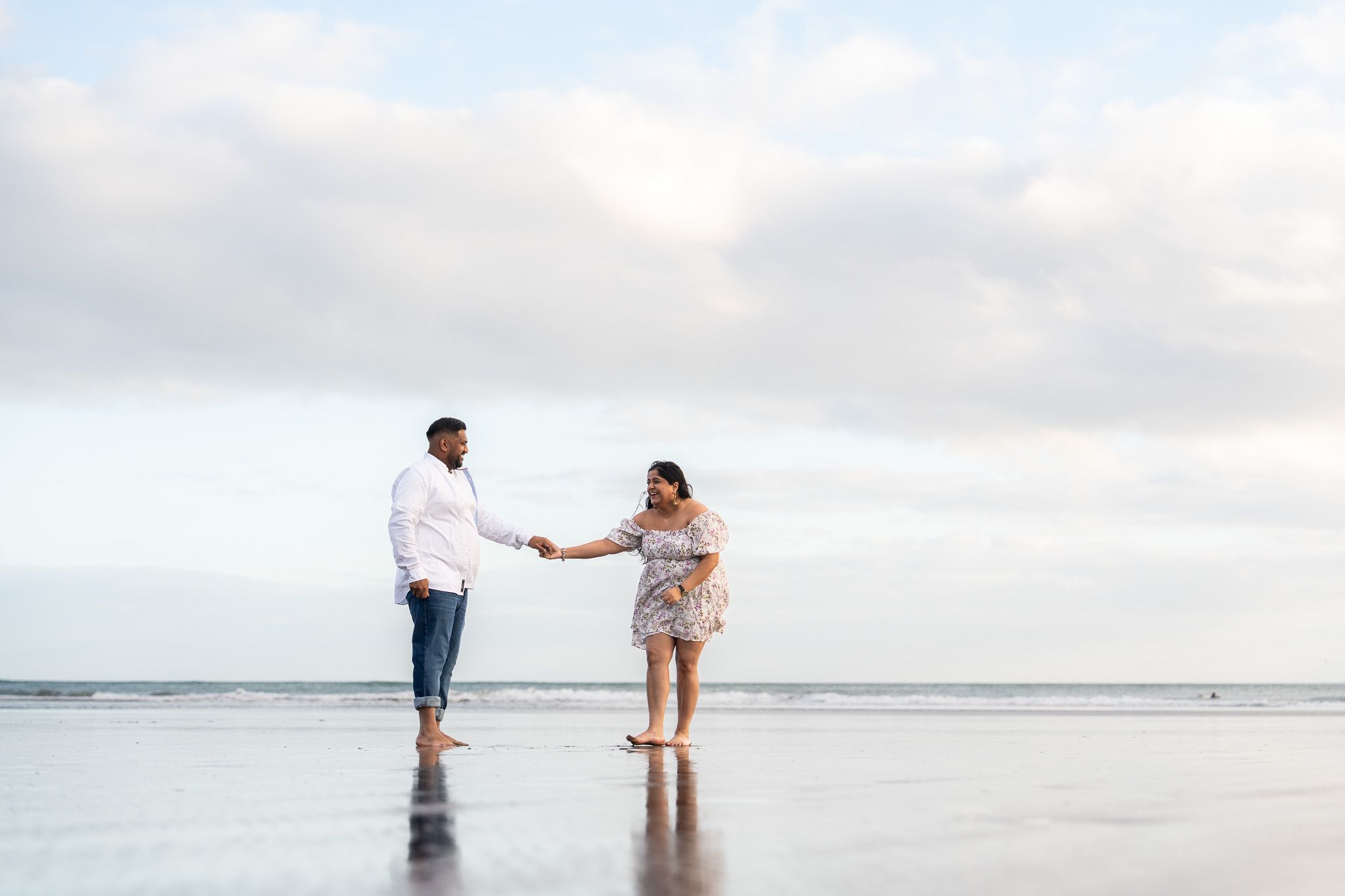 Joyful couple laughing and holding hands during an engagement session at Muriwai Beach in Auckland, surrounded by sea, clouds and soft light