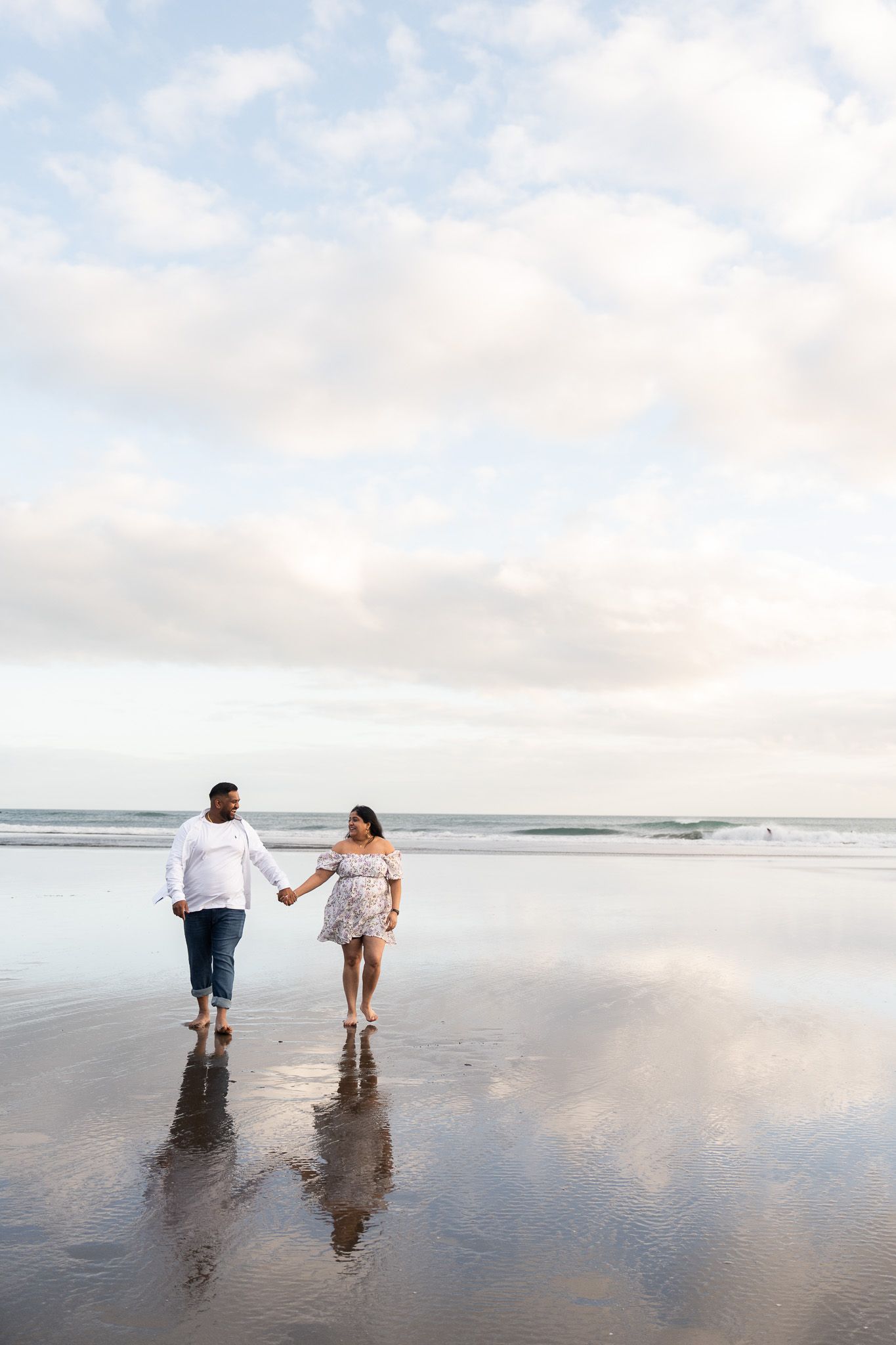 Barefoot couple walking hand-in-hand through shallow reflective water at Muriwai Beach in Auckland during their candid beach engagement session