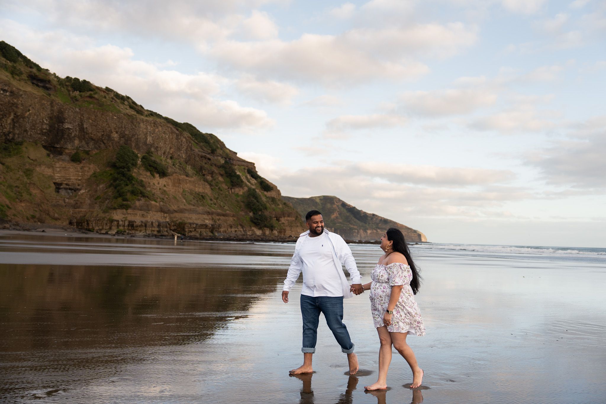 Couple walking across the shoreline at Muriwai Beach in Auckland, laughing together with dramatic cliffs in the background during golden hour