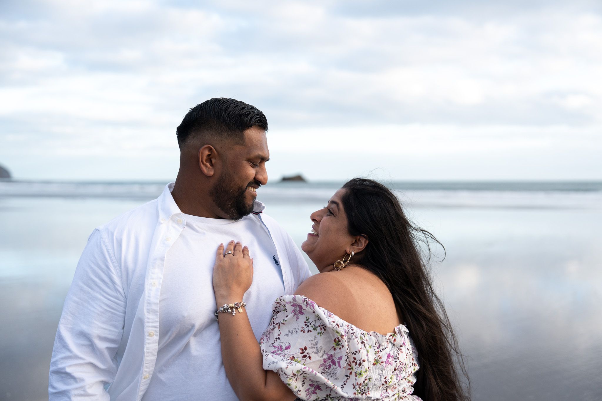 Playful and romantic moment as couple lock eyes and smile on Muriwai Beach in Auckland, captured during a candid couple photography shoot