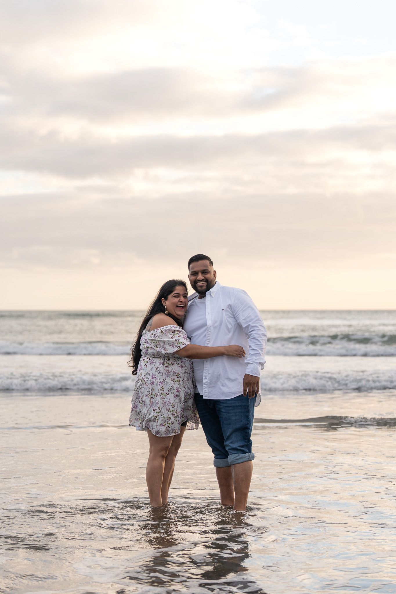 Couple laughing and hugging while standing ankle-deep in the ocean at Muriwai Beach in Auckland, taken during an engagement photography session