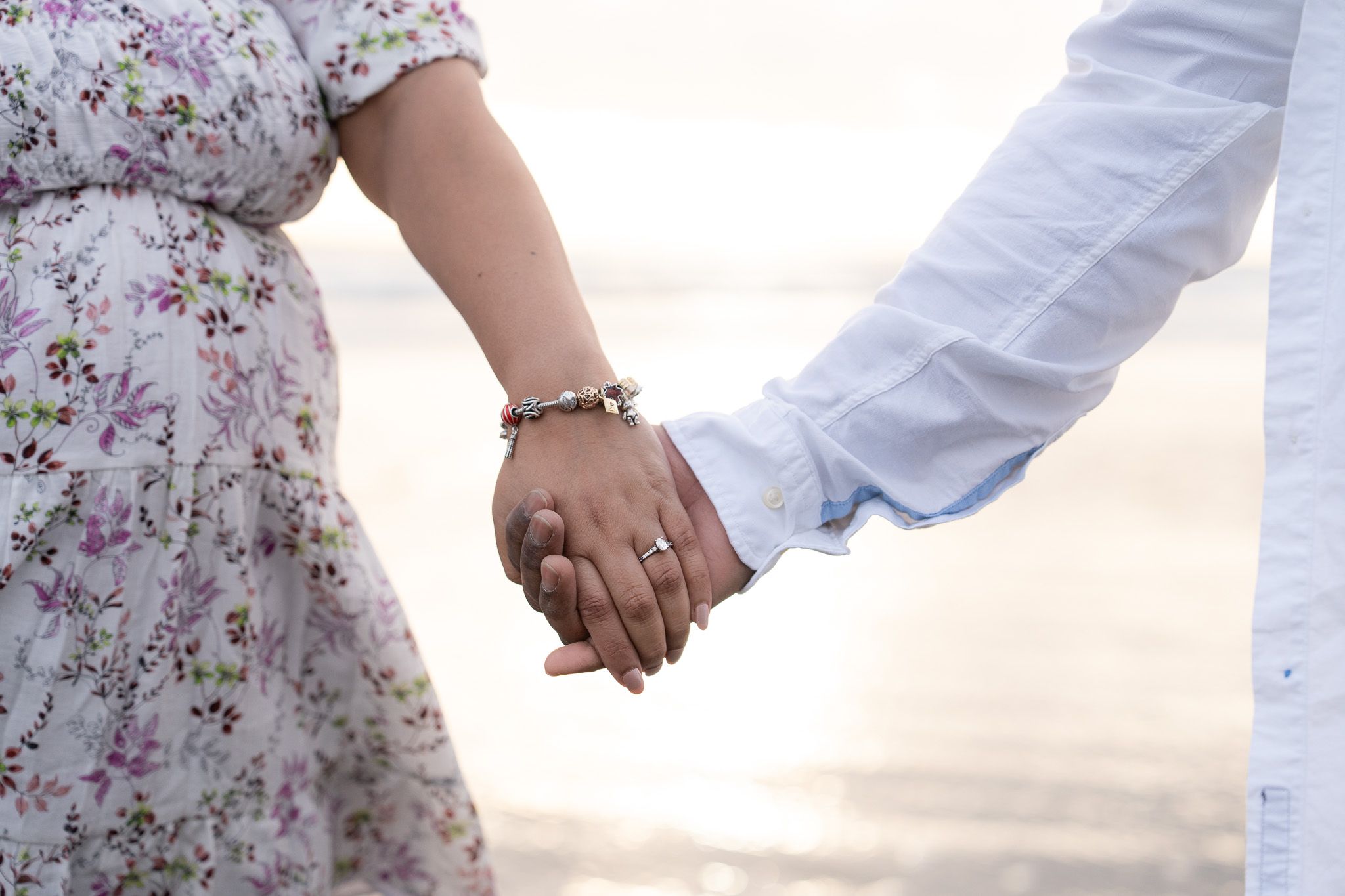 Engagement ring and charm bracelet in focus as couple hold hands by the water during a candid couple photography session at Muriwai Beach, Auckland