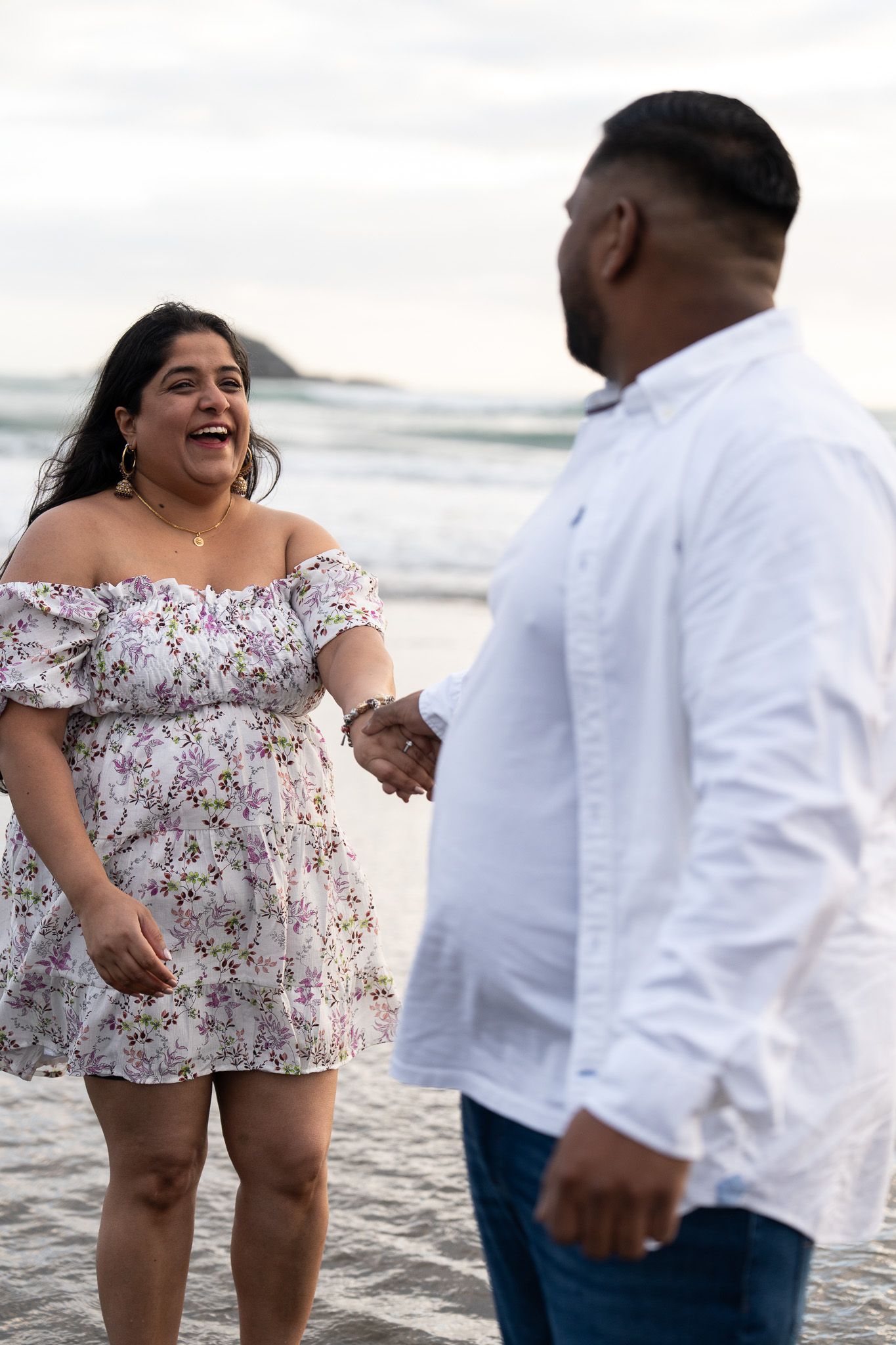 Candid moment of a woman laughing while holding hands with her partner during a beach couple shoot at Muriwai Beach in Auckland