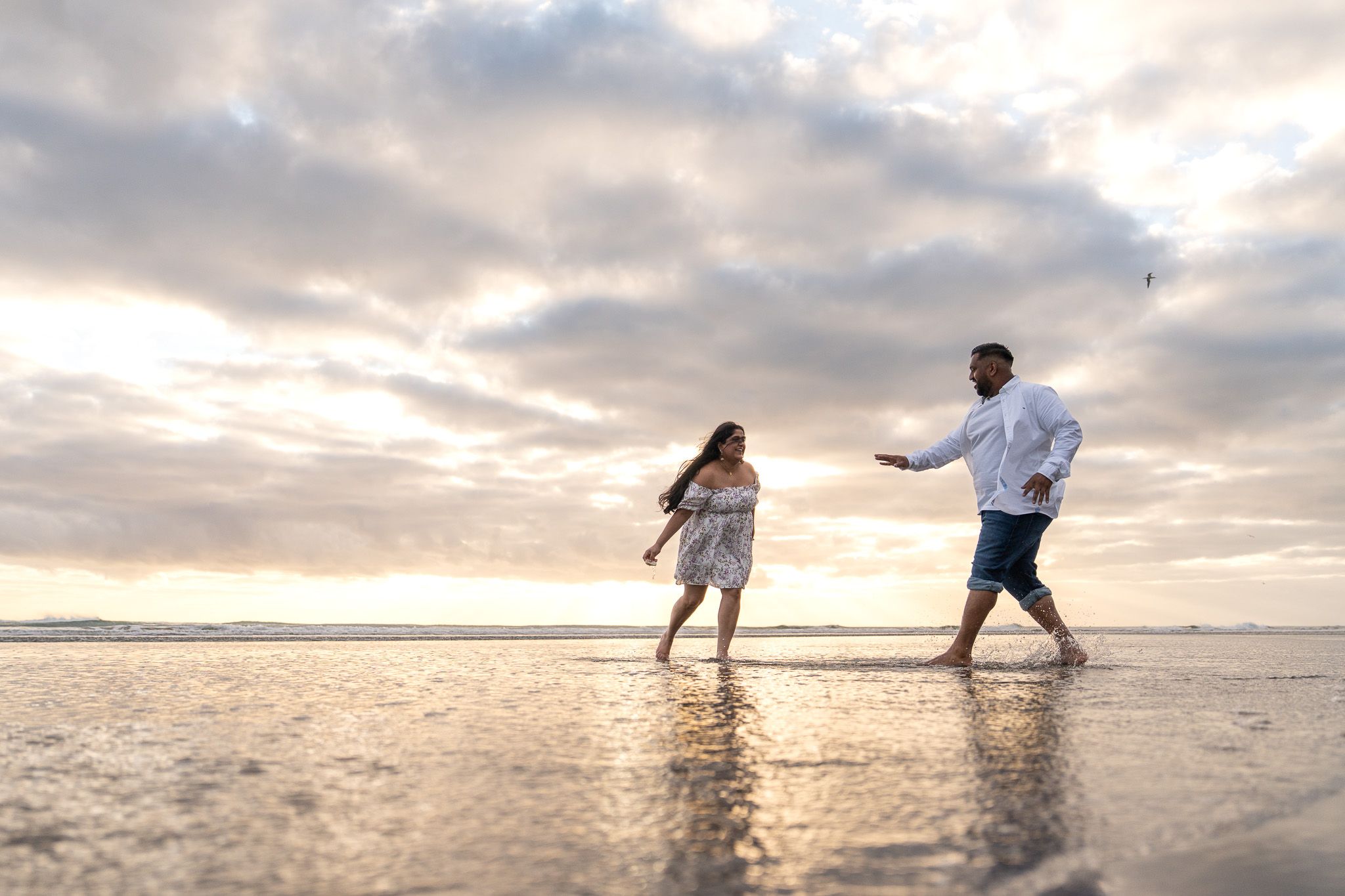 Romantic moment of couple splashing through shallow water at Muriwai Beach in Auckland during sunset, captured during playful couple photography