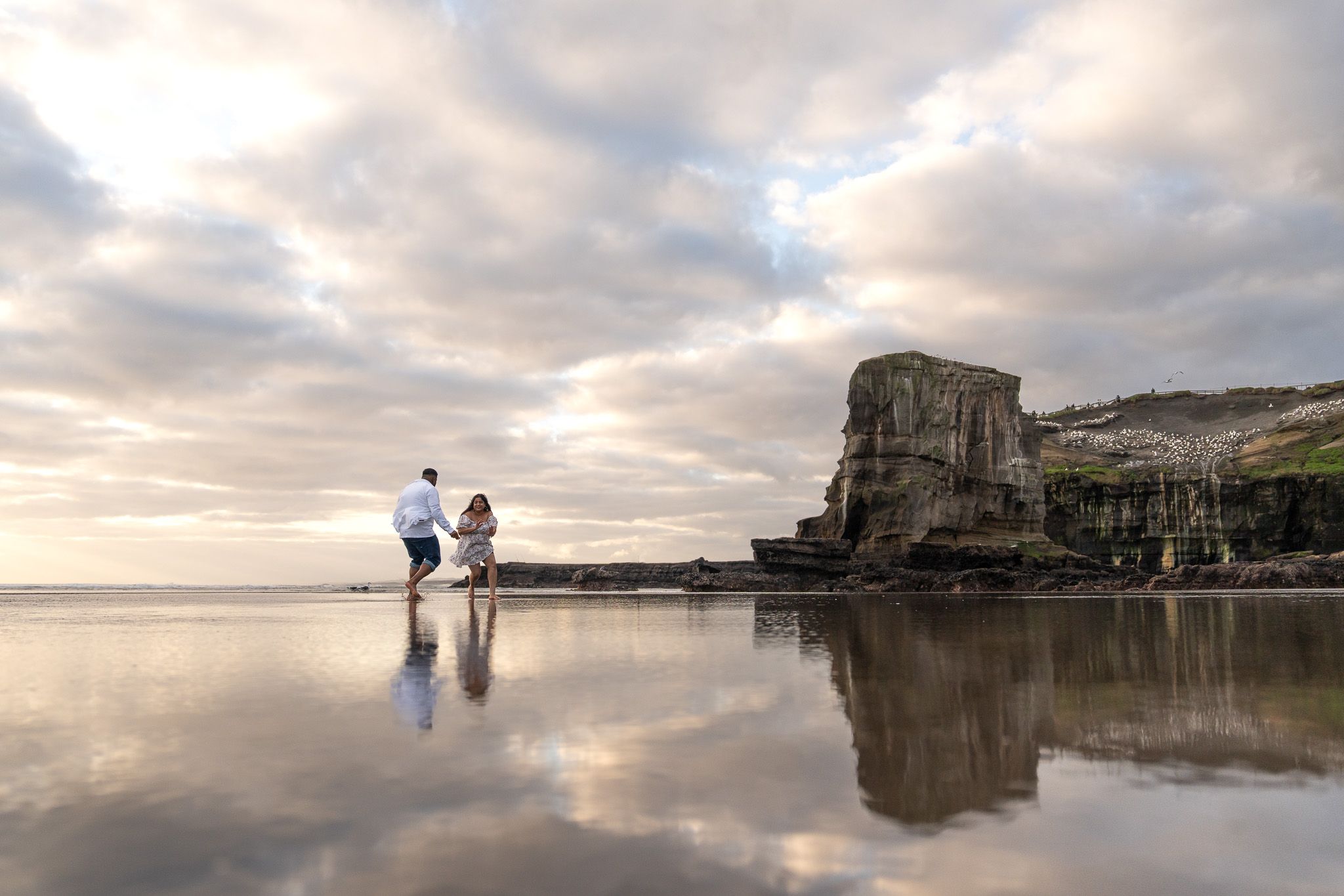 A couple stands in the soft golden sand at Muriwai Beach, gently holding each other with a calm ocean stretching out behind them under soft light
