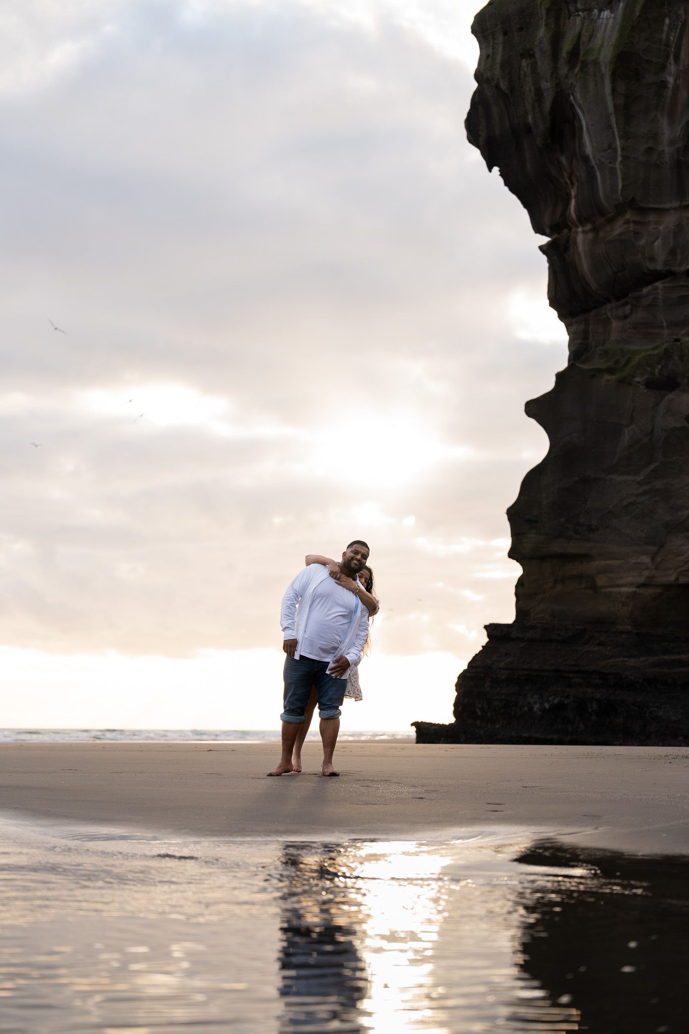 Smiling woman hugging her partner from behind as they pose in front of the dramatic cliffs at Muriwai Beach during a fun couple photography session in Auckland
