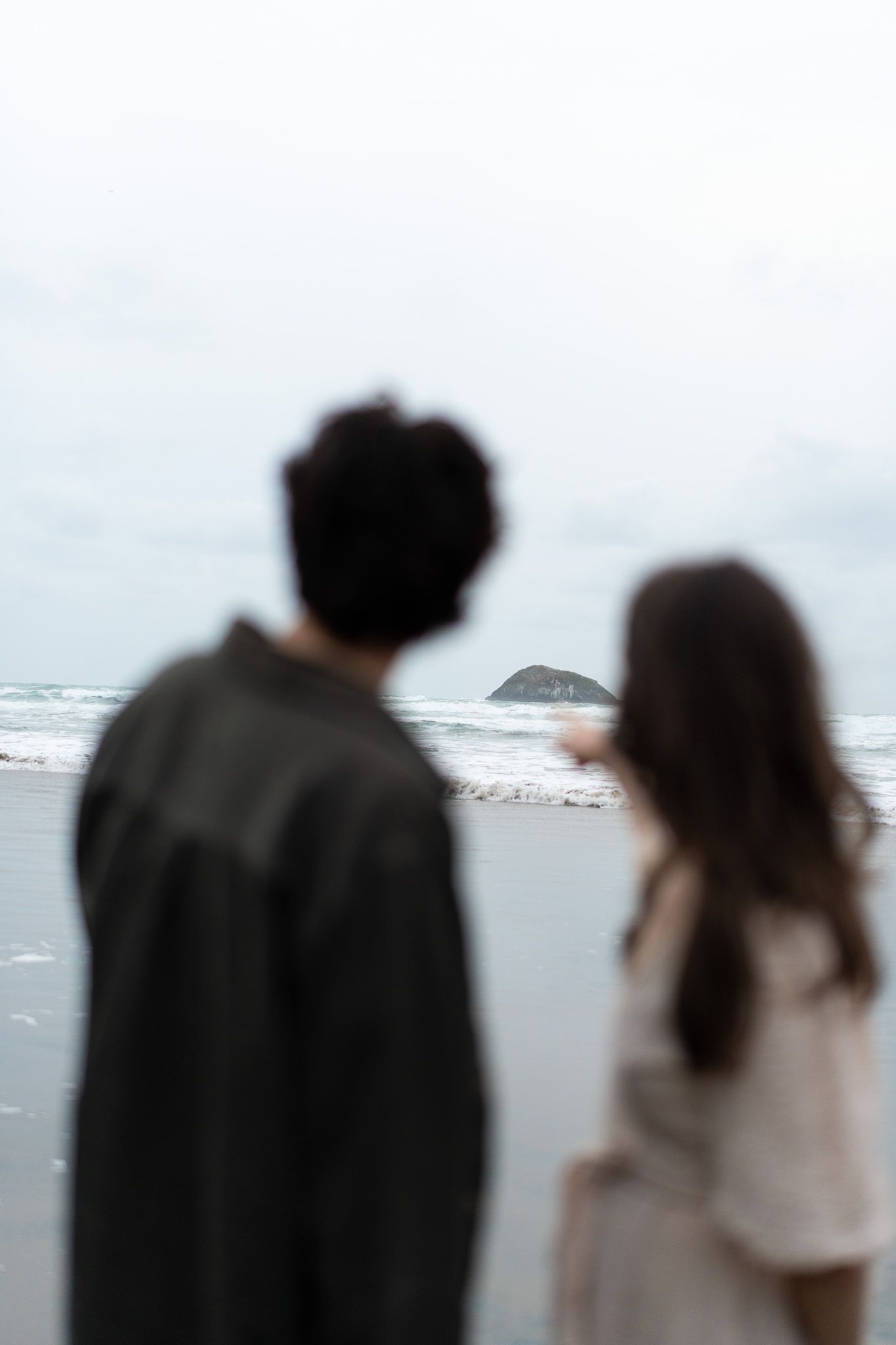 Couple standing on the sand looking toward the ocean as the woman points to the island in the distance during a beach photoshoot in Auckland