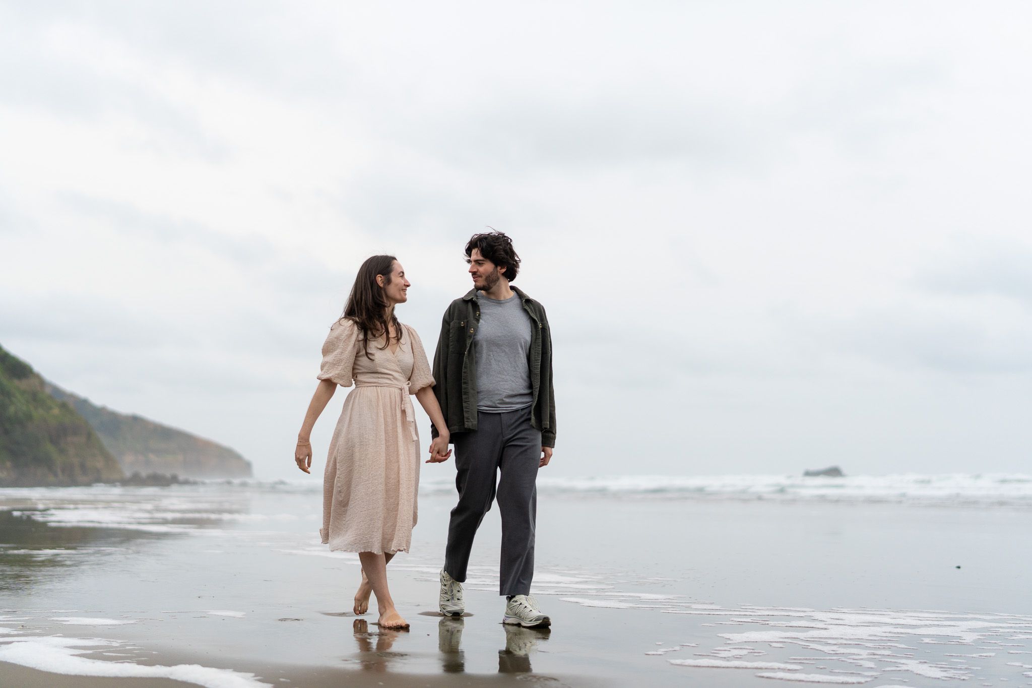 Couple walking hand in hand along the shoreline sharing a quiet smile during their relaxed couple photography session at Muriwai Beach in Auckland