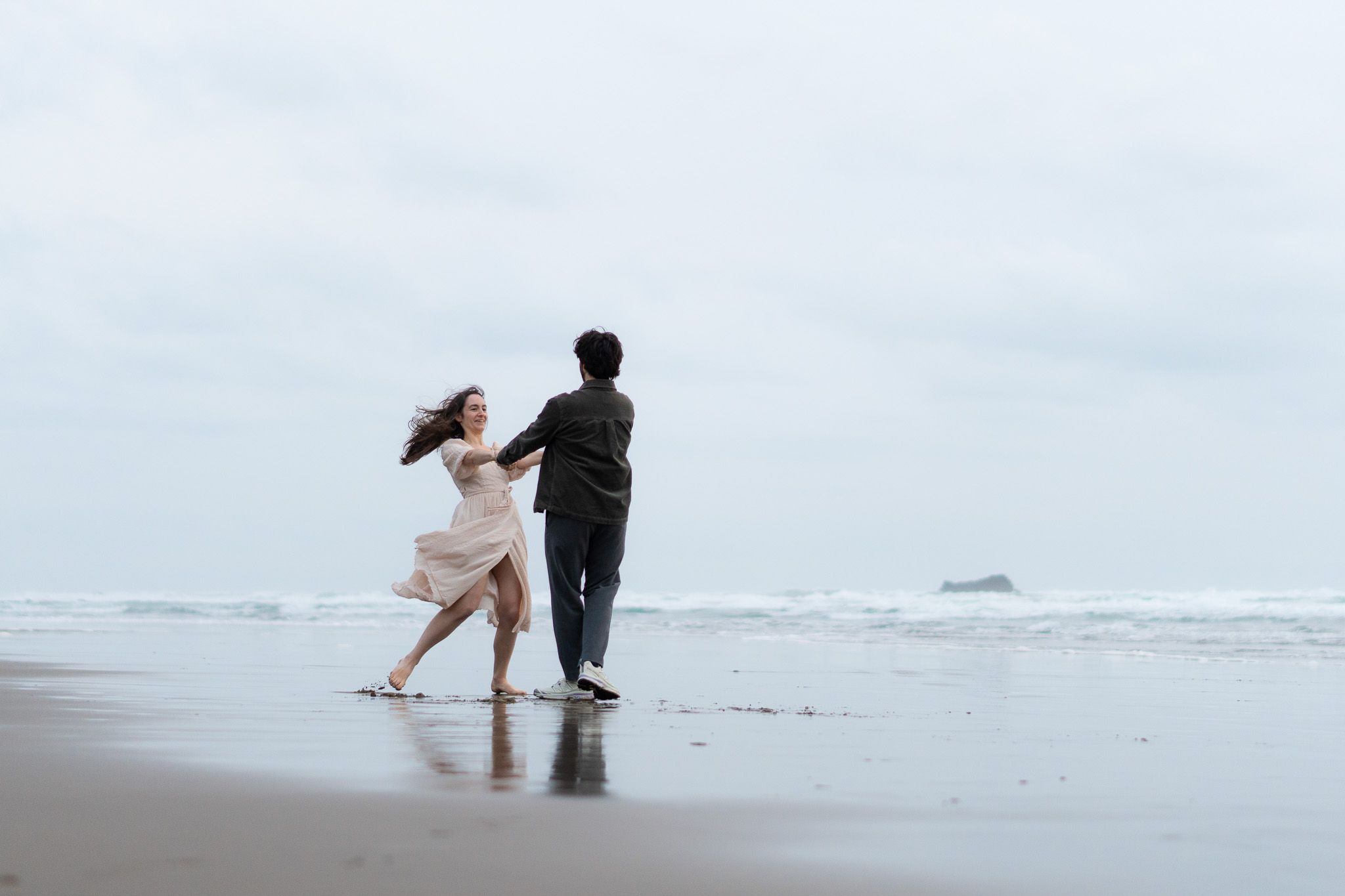 Couple holding hands and spinning joyfully on the shoreline with flowing dress and gentle waves at Muriwai Beach in Auckland