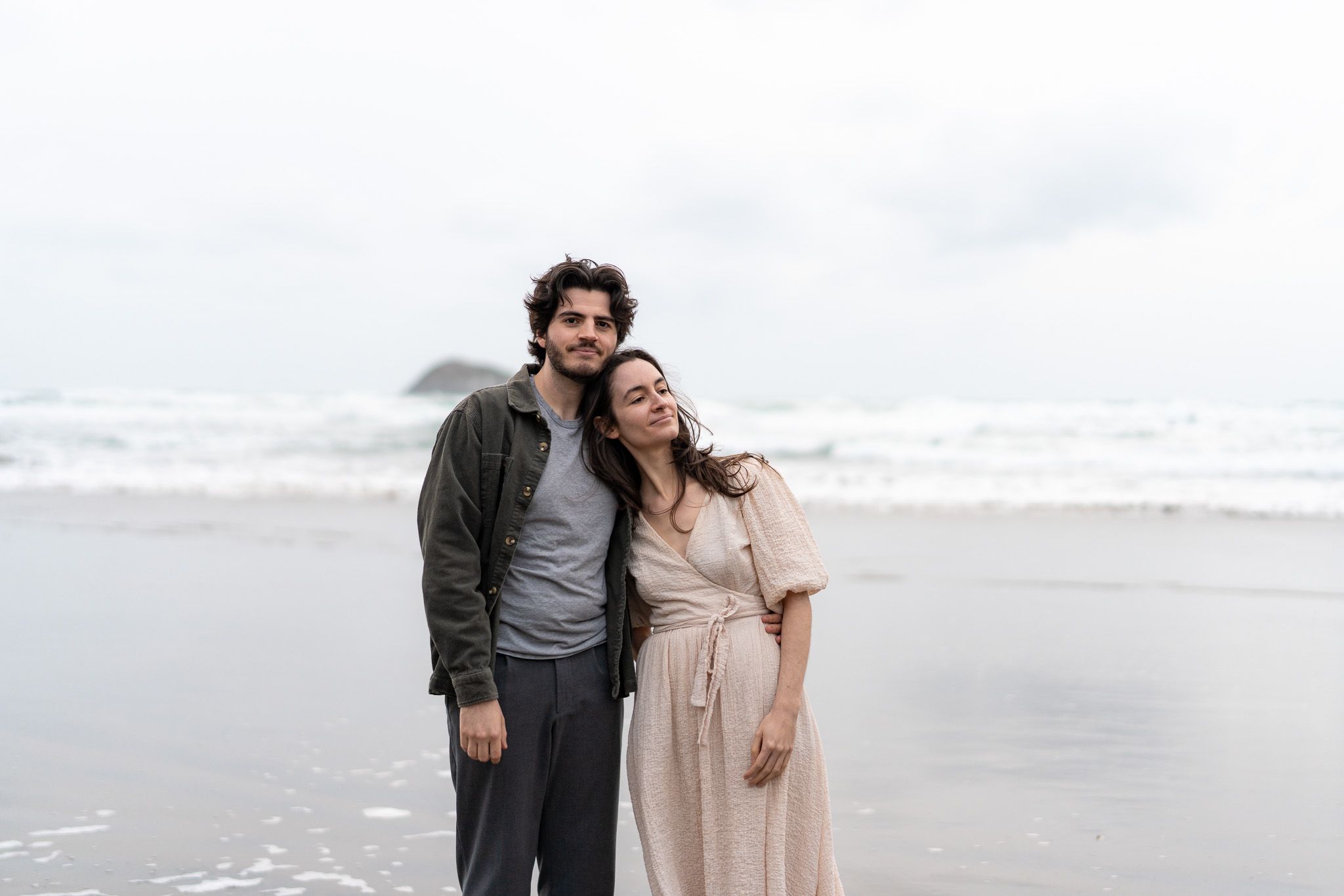 Couple embracing by the ocean with gentle wind and overcast skies during a romantic beach photography session in Auckland