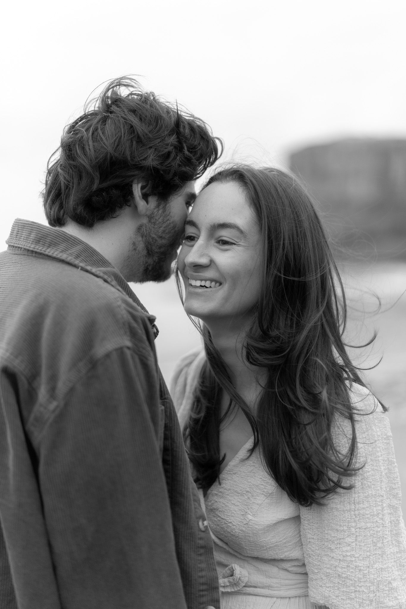 Couple sharing a quiet moment laughing together with the rugged coastal cliffs and ocean waves behind them at Muriwai Beach in Auckland