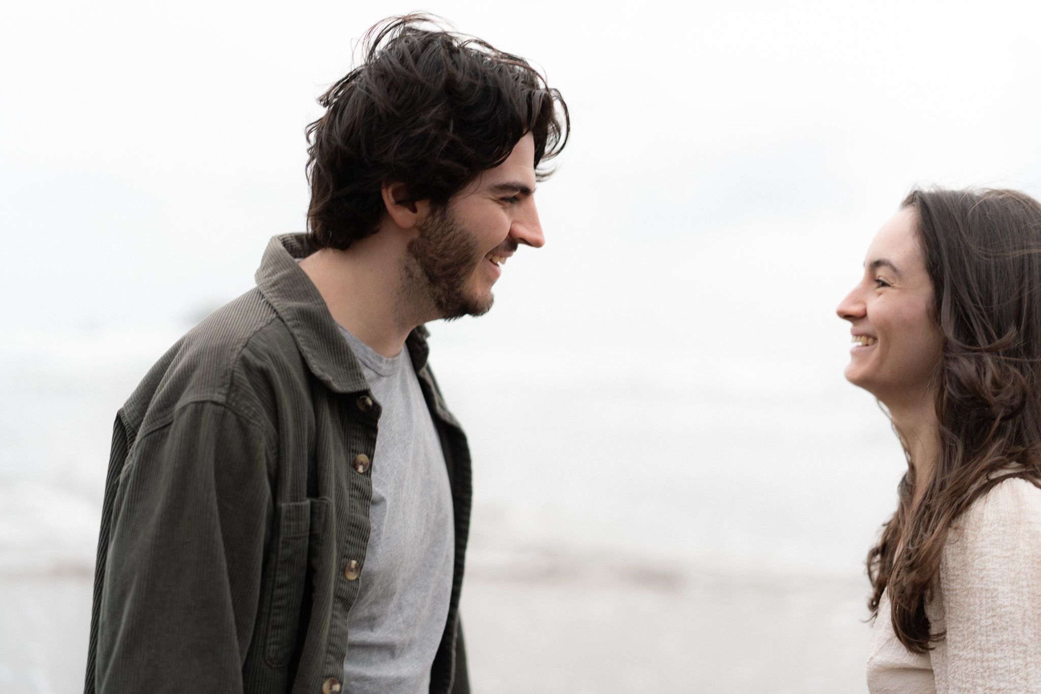 Couple smiling at each other on the beach with soft natural light and ocean breeze during their relaxed couple photoshoot in Auckland