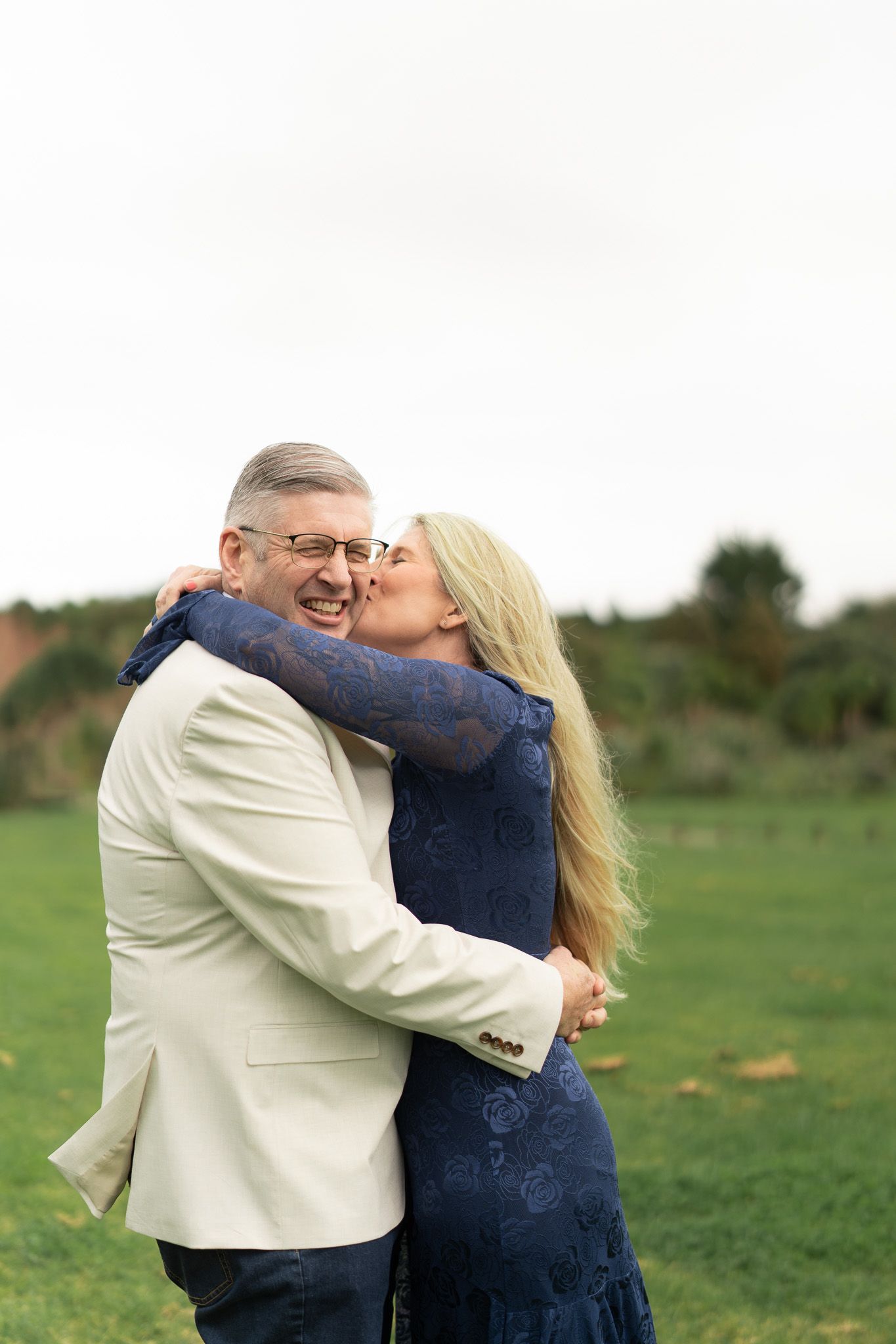 Bride kissing the groom's cheek as he laughs, embracing on the grass