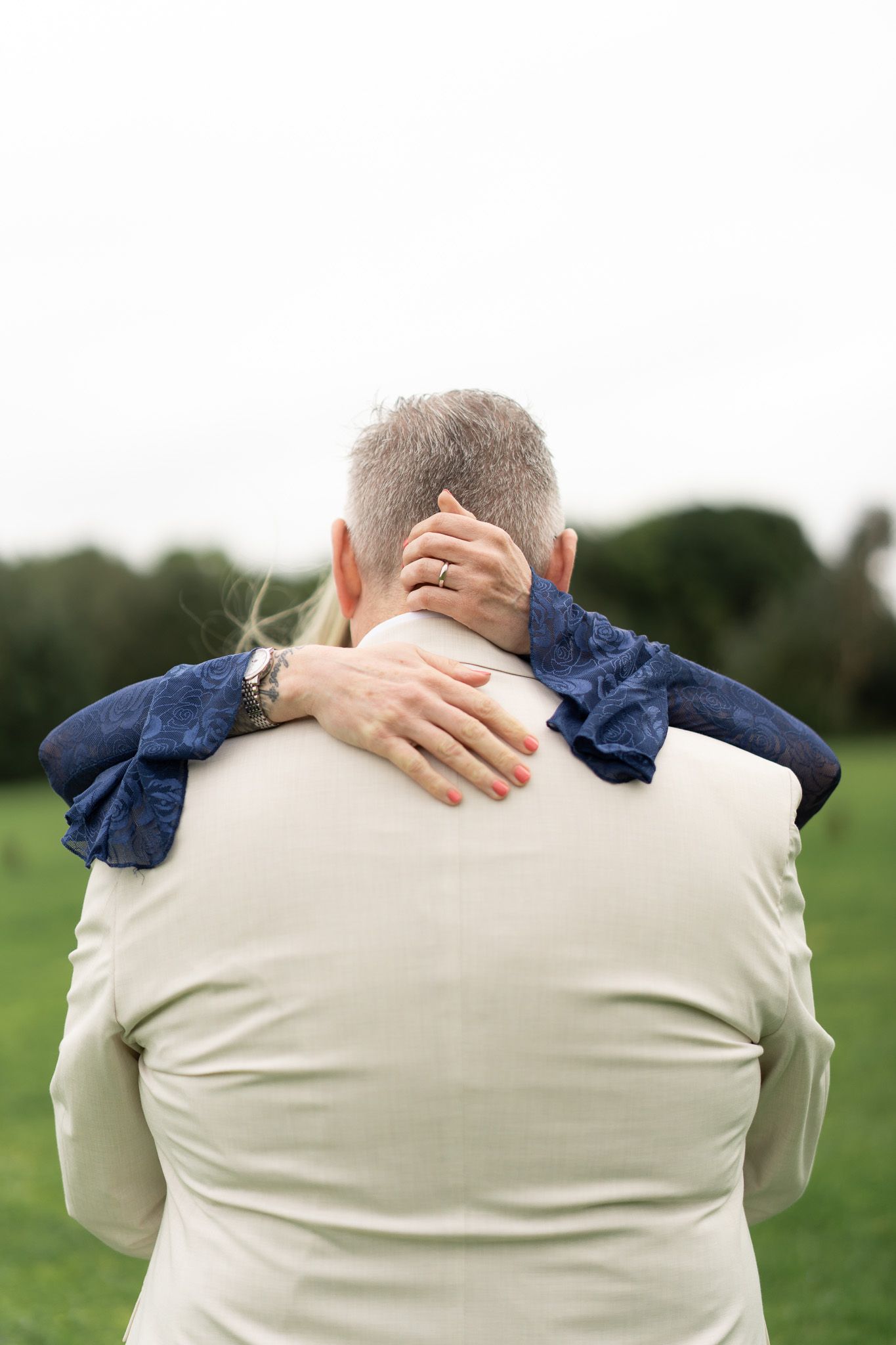 Close-up of the bride's arms around the groom's neck, her wedding ring visible, on the grass