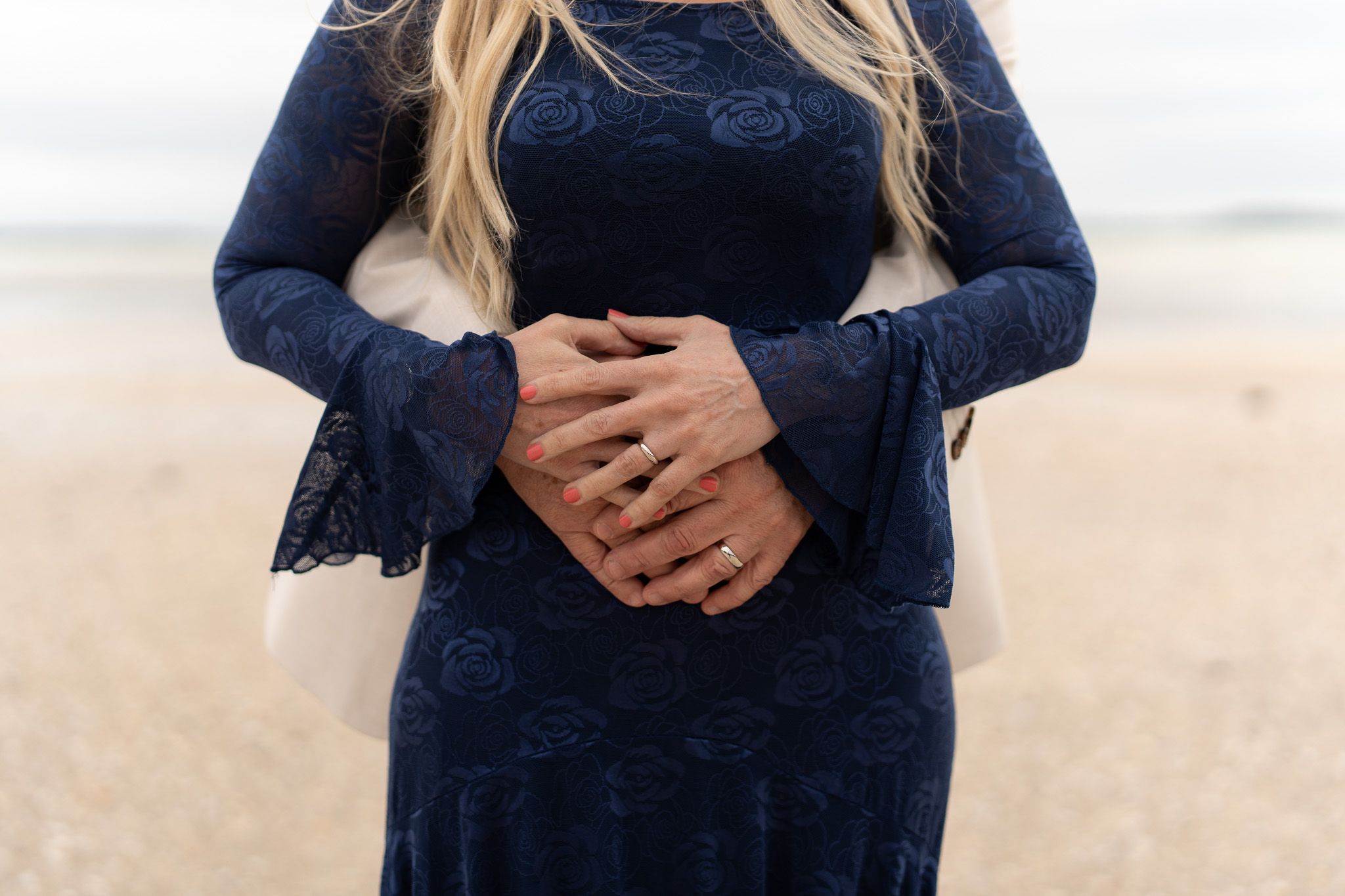 Close-up of the bride and groom's clasped hands resting against her lace dress on the beach