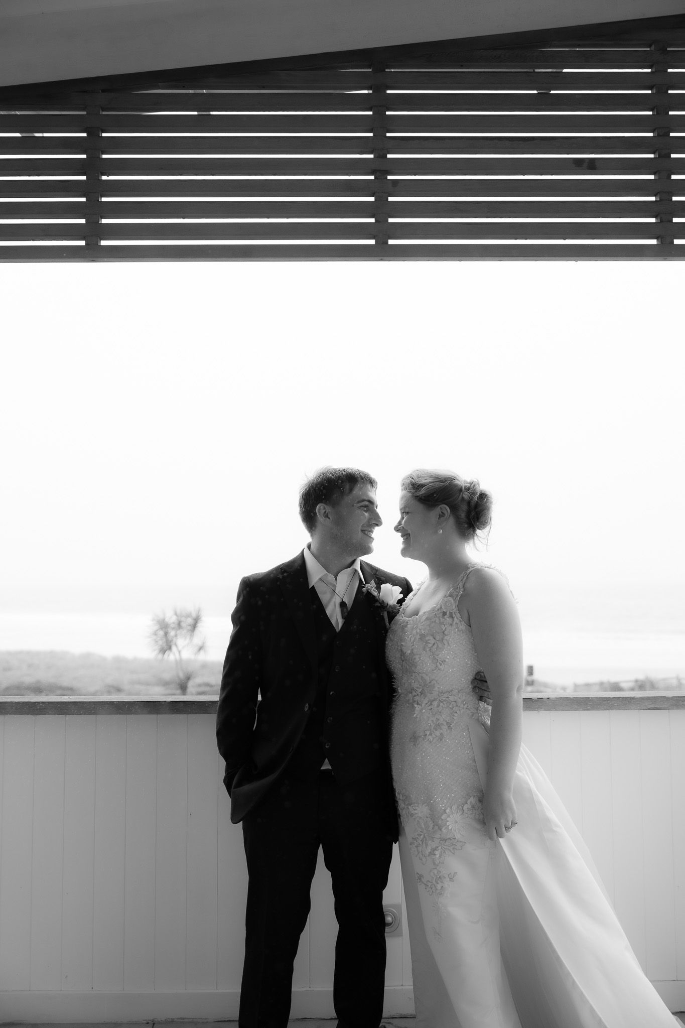 Black and white close-up of bride and groom embracing under a pergola with the beach visible behind them