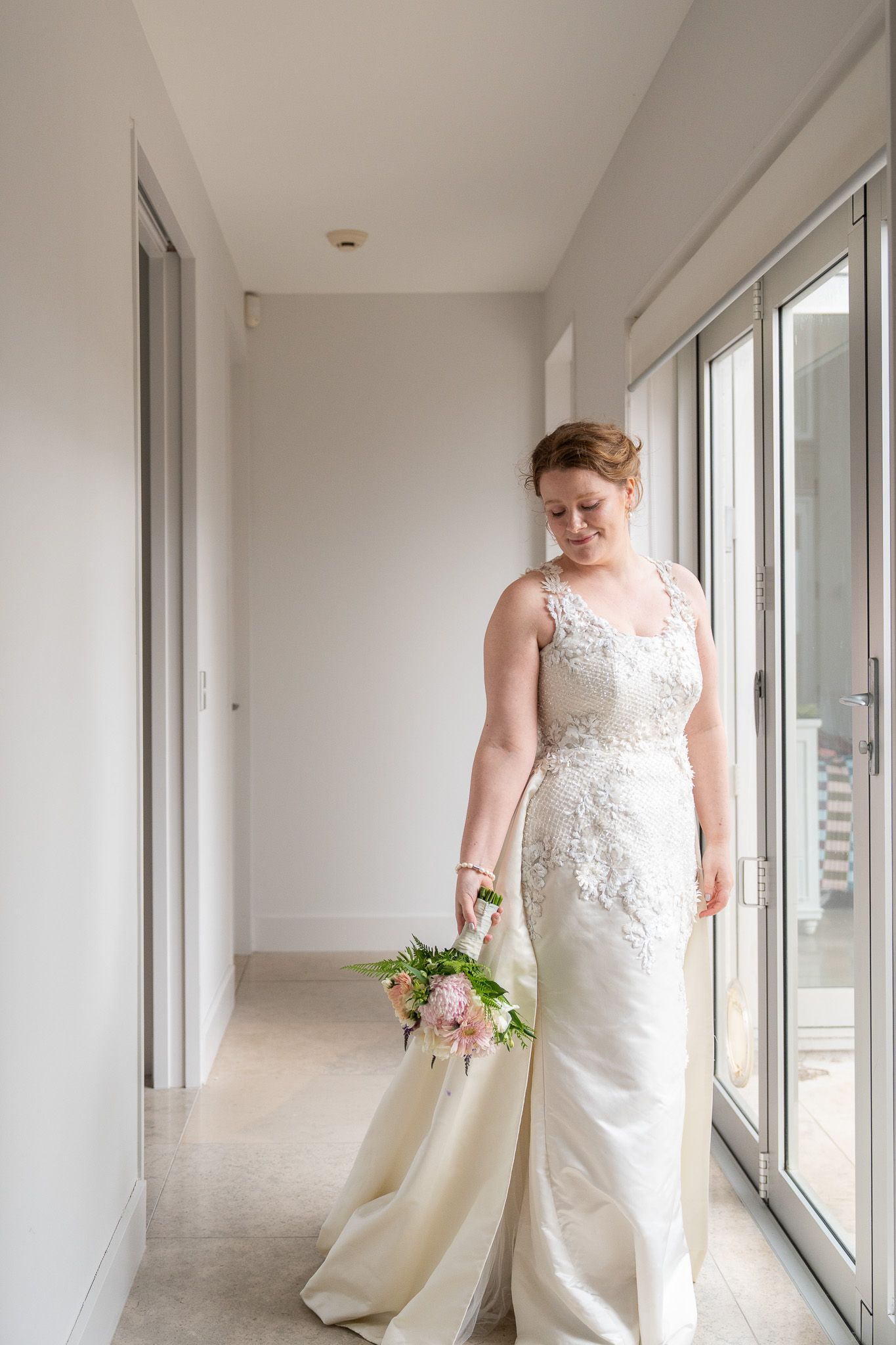 Bride alone in a sunlit hallway, looking down at her bouquet