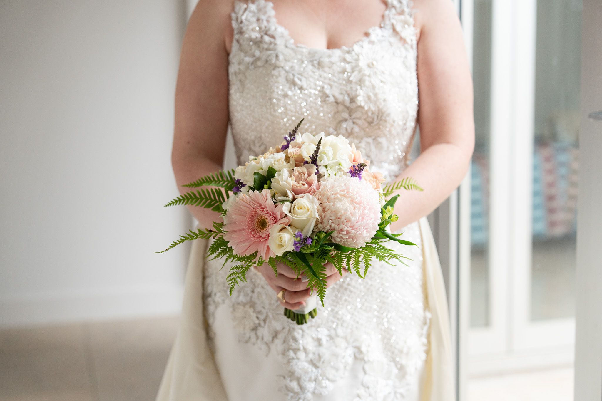 Close-up of a pastel bouquet of roses and gerberas held against a beaded lace wedding gown