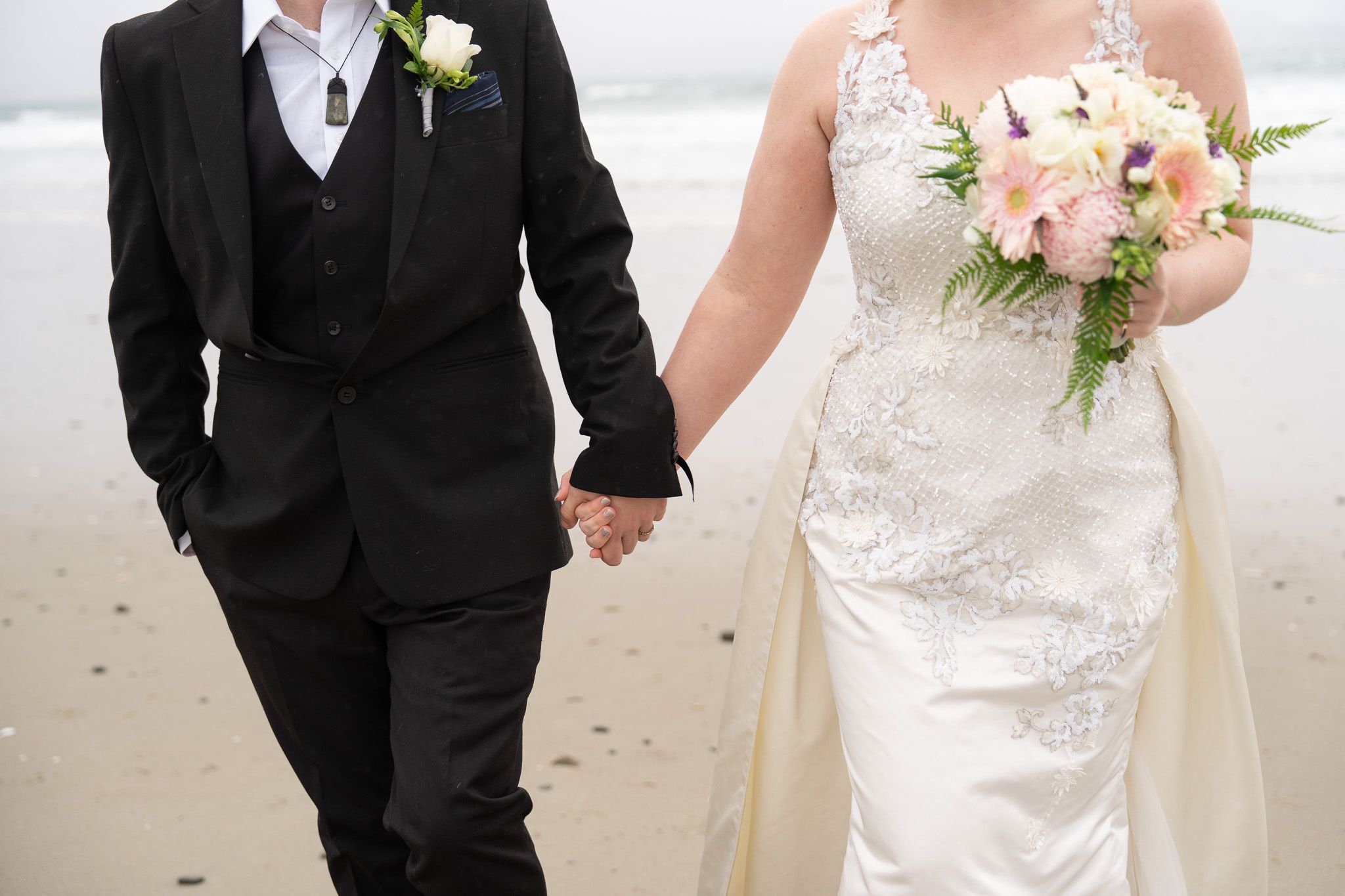 Close-up of bride and groom holding hands on the beach, her bouquet held between them