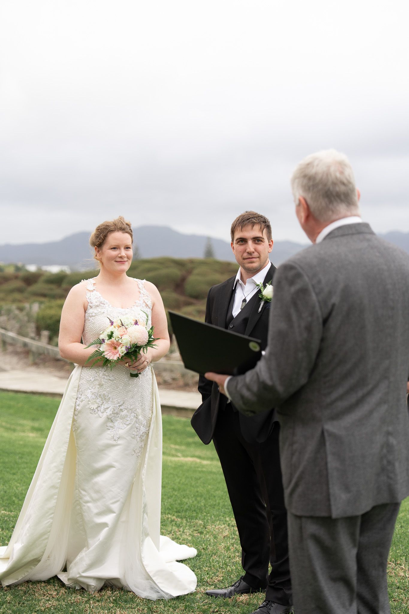 Bride and groom exchanging vows on the lawn at their Omaha holiday home as the celebrant reads, hills and coastline behind them