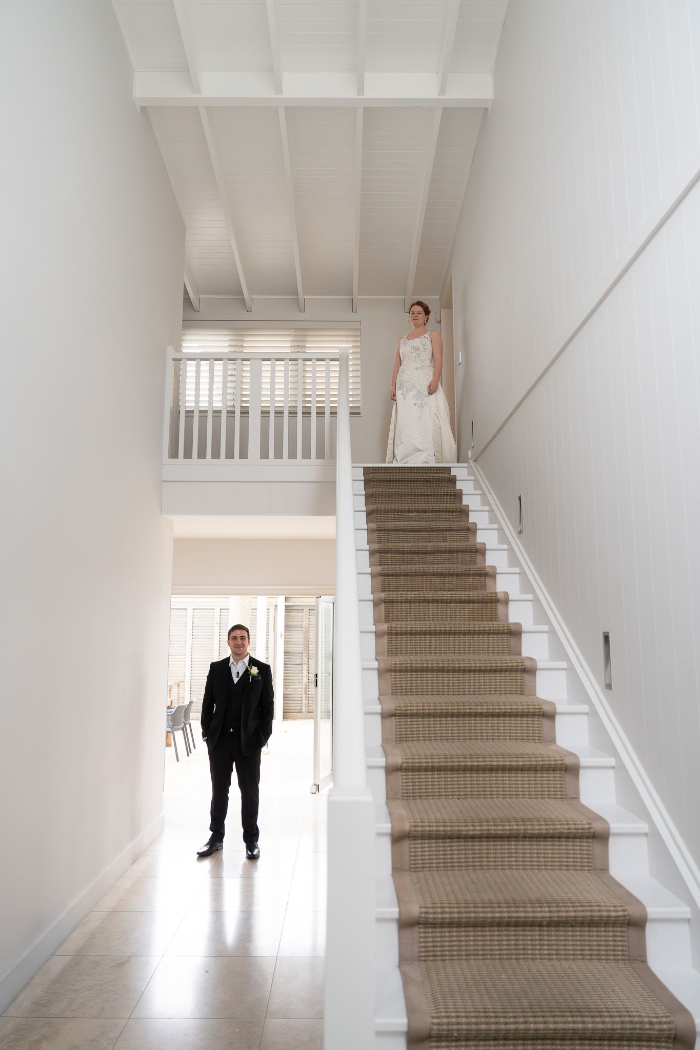 Groom waiting at the bottom of a staircase while the bride looks down from the landing above