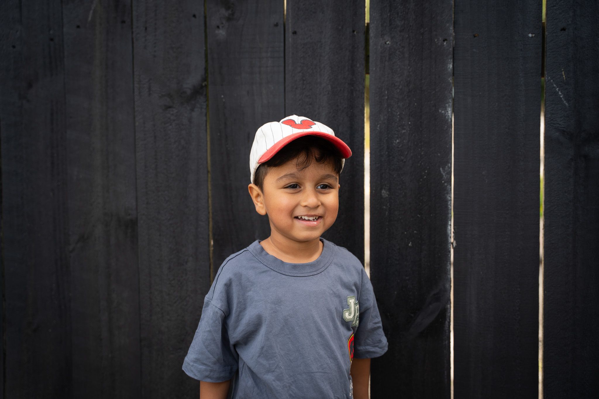 Smiling young boy wearing a grey t-shirt and red-white cap standing against a dark wooden fence during a relaxed family photoshoot in Auckland